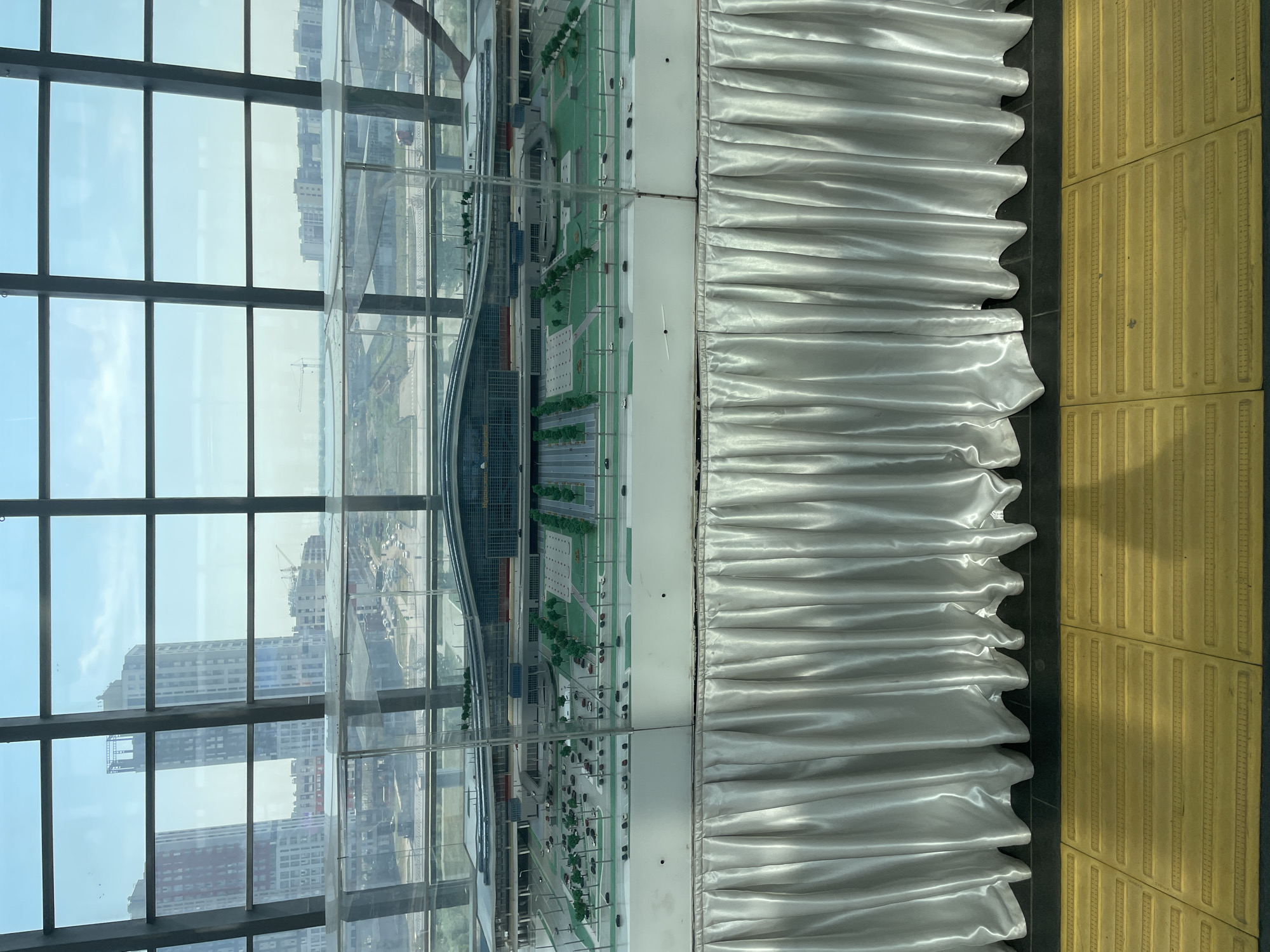 The image shows a miniature architectural model of a large building, possibly a transportation hub or airport, displayed behind a clear protective casing. The model features detailed landscaping with miniature trees, roads, and parked cars, suggesting a functional urban environment. The building itself has a modern, sprawling design with a curved, reflective roof. The model is elevated and placed on a white base covered by a white satin tablecloth with cascading folds. In the background, through a large glass window pane divided by dark metal frames, a cityscape is visible with contemporary high-rise buildings and cranes, indicating ongoing construction. The sky is bright and appears to be daytime. In the foreground, a textured yellow tactile paving surface is visible, common in public spaces for accessibility. This scene is located in Astana, Kazakhstan, as indicated by the presence of the city's characteristic modern architecture and the specific design of the building model, which resembles the Nur-Sultan Nazarbayev International Airport.