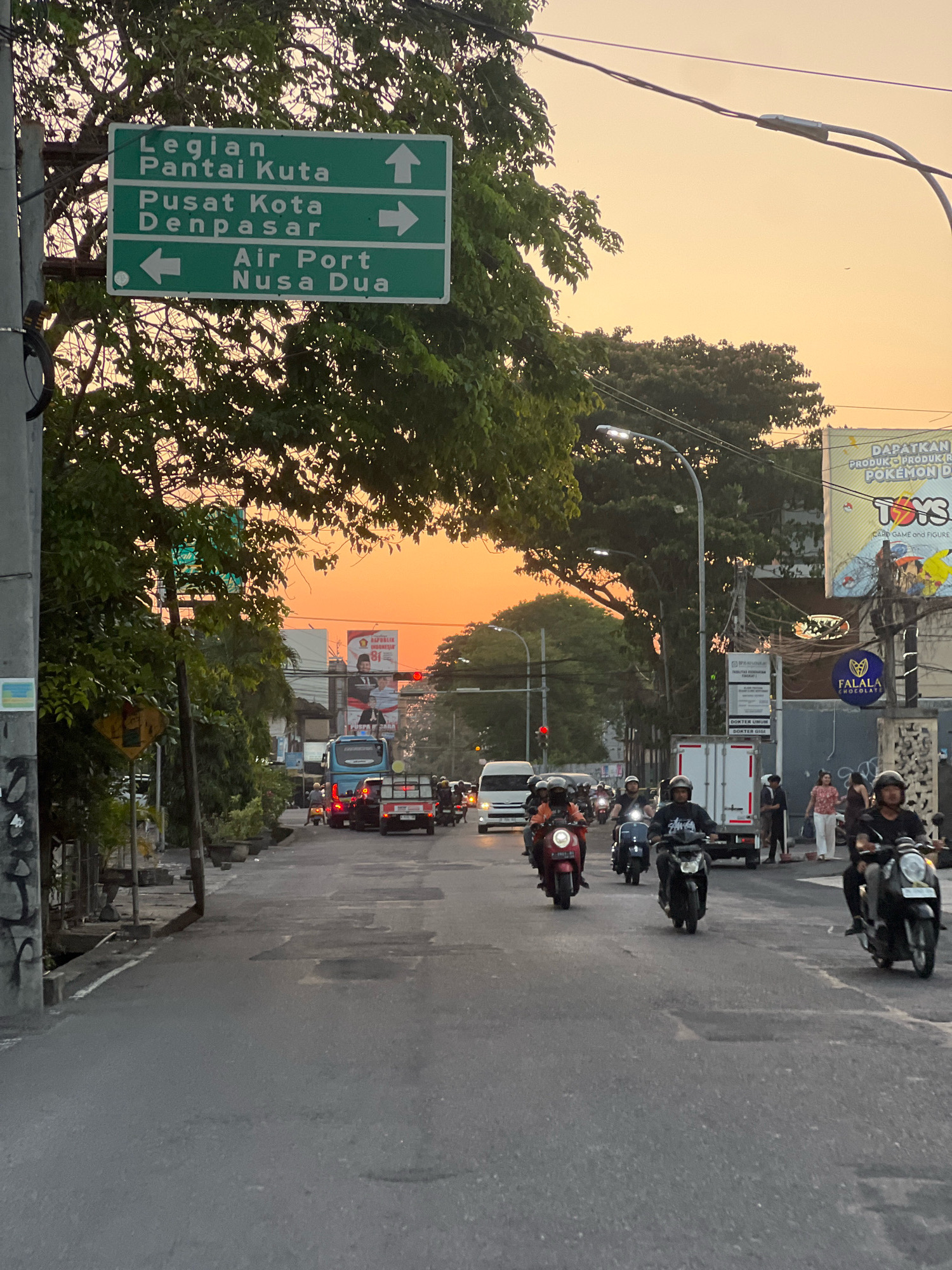 The image depicts a street scene in Kuta, Indonesia, at dusk, with a clear orange sky visible. A prominent green directional sign is mounted on a pole, indicating directions to "Legian Pantai Kuta" (up arrow), "Pusat Kota Denpasar" (right arrow), and "Air Port Nusa Dua" (left arrow). The street is paved and shows signs of wear and tear. Traffic is present, with several motorbikes carrying individuals riding in the same direction as the camera. Some individuals are wearing helmets, while others are not. In the background, there are trees, buildings, streetlights, and various advertisements on the sides of structures. One advertisement appears to be for "Toys" and possibly "Pokemon." A blue truck and other vehicles are also visible in the distance. The overall atmosphere is that of a busy urban environment at the end of the day.