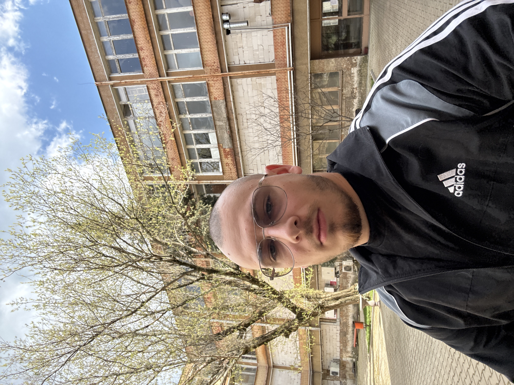A photo of a man wearing sunglasses and an Adidas tracksuit stands in front of a brick building and a tree with budding leaves. The building has multiple windows with visible window frames, and some sections appear worn or aged. The street is paved with interlocking stones. The sky is partially visible through the tree branches, showing patches of blue and some cloud cover. Based on the architectural style and the presence of similar structures, the location is likely in Stara Zagora, Bulgaria.