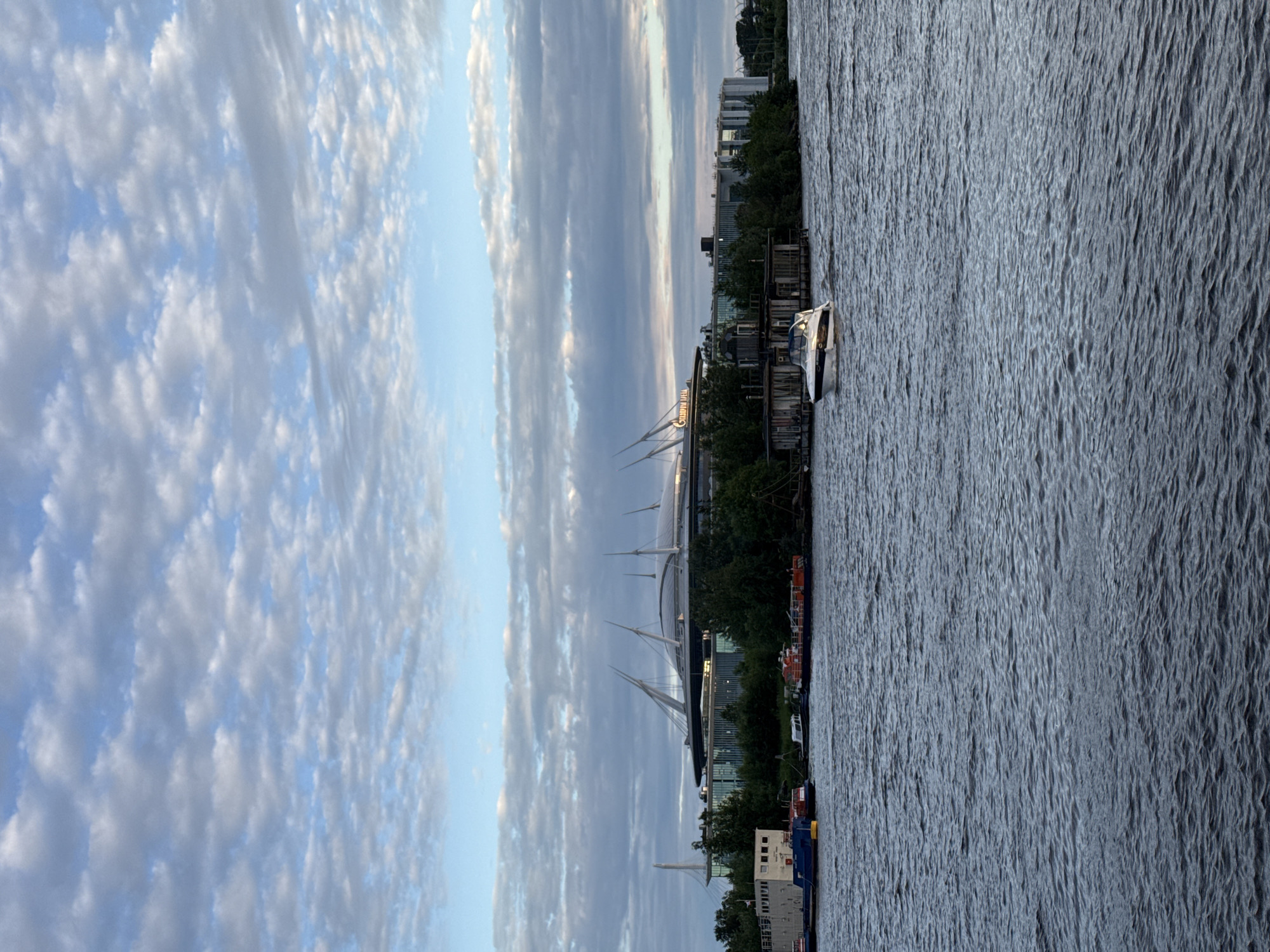 The image depicts a wide, dark blue body of water with gentle waves in the foreground, likely a river or bay. A white motorboat with a dark stripe and illuminated cabin is moving from right to left across the water, creating a wake. In the mid-ground, a tree-lined embankment is visible, with several small, rustic structures and a white barge with blue and red accents docked along the shore. Behind the embankment, a large, modern stadium with a distinctive curved roof supported by slender white masts dominates the skyline. The stadium's name, "GAZPROM ARENA," is illuminated in large letters on its side. The sky above is a soft blue, filled with scattered white and gray clouds, suggesting a partly cloudy day, possibly at dusk. The overall scene captures a view of the Gazprom Arena stadium in Saint Petersburg, Russia, from a body of water.