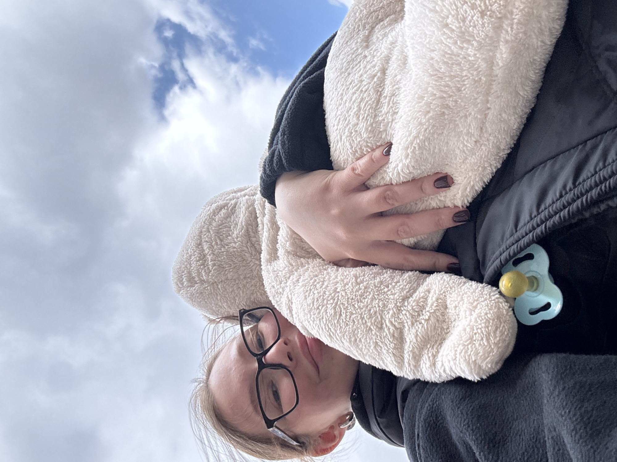 An upward-angle shot captures a person, likely an adult female, holding what appears to be a baby or small child wrapped in a fluffy, light-colored garment or blanket. The adult's head is inverted in the frame, wearing black-rimmed glasses and looking down towards the camera with a neutral expression. Their light hair is partially visible. One hand, adorned with dark nail polish, rests on the soft, cream-colored material covering the child. The adult is dressed in dark-colored, possibly fleece, clothing, topped with a black jacket or vest. A light blue pacifier with a yellow nipple is tucked into the adult's clothing on the right side of the frame, near the fluffy garment, reinforcing the presence of a baby. The background features a bright blue sky dotted with scattered white clouds, suggesting a daytime setting with pleasant weather. The image composition implies it was taken from a low vantage point, possibly from the perspective of the child or someone positioned below. There is no visible text in the image. The location context provided is Chaikovsky, Russia.