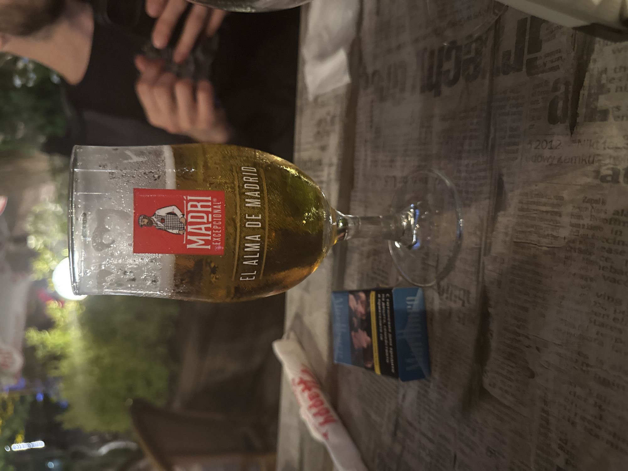 The image depicts a casual evening scene, likely set at an outdoor bar or restaurant in Burgas, Bulgaria. A glass of cold Madri Excepcional beer, with condensation on its surface, is prominently featured. Its red label displays a man in a hat and the text "MADRI EXCEPCIONAL®" and "EL ALMA DE MADRID" (The Soul of Madrid).
Beside the beer glass, a blue and black cigarette pack is visible, featuring a health warning in Bulgarian Cyrillic script, such as "Cърцетови напълно ето. цигари," consistent with the location context. A white wrapper, partially showing the word "Happy," lies nearby.
The table surface is covered with a decorative print resembling old newspaper pages. This print includes fragmented texts in multiple languages, such as English ("kers. A", "arlech as", "Stares"), Bulgarian Cyrillic ("на децата, семейството", "Статииевия дала вредите"), and possibly Polish ("2012. Nikt 10", "edowy zamku").
In the upper left quadrant, a person is partially visible, showing their beard and hands holding an indistinct object. The background is softly blurred with hints of green foliage and out-of-focus lights, suggesting an outdoor setting during the evening or night. No specific emotions are discernible from the obscured person, and no particular weather conditions are evident.