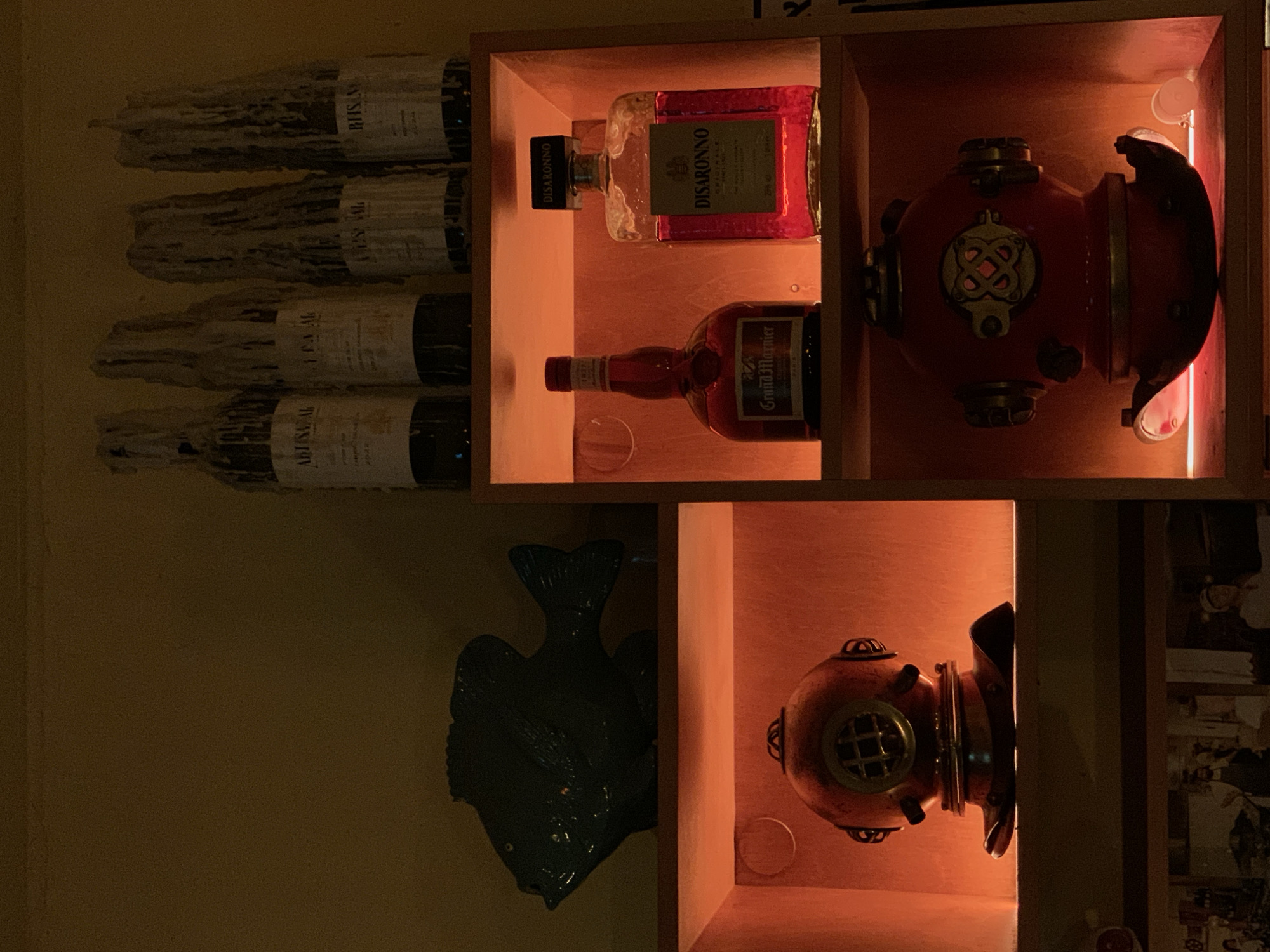 This image depicts an interior display from Abusal Restaurant in Jaffa, Tel-Aviv, Israel, characterized by dim, warm, localized lighting. The scene features dark wooden shelving against a muted olive-green or dark yellow wall, showcasing various objects and liquor bottles.
On the topmost unlit shelf, four dark glass wine bottles stand upright, each heavily encrusted with dried white candle wax. Visible labels include "ABUSAL" on the leftmost bottle, followed by "LE'A MAL", "LES' SAL", and "BHSN" on the subsequent bottles to the right.
Below this, a dark, possibly ceramic or resin, fish figurine is positioned horizontally on a flat, unlit wooden shelf. Its surface appears dark green or black in the ambient light.
Adjacent to the fish, a vertical stack of three recessed wooden cubbies is illuminated by a uniform warm, orange-red light source. The top cubby displays two liquor bottles: on the left, a bottle of Grand Marnier liqueur with a red-brown liquid and a label reading "Grand Marnier PARIS"; on the right, a clear bottle of Disaronno Originale containing a bright reddish-pink liquid.
The middle cubby on the left features a decorative diving helmet, rusty red-orange in color with brass-colored accents, including a circular viewport with a grid pattern. The lowest cubby on the right contains a similar rusty red-orange diving helmet, distinguished by an ornate brass-colored plate on its front.
Below the illuminated shelves, the top edges of several framed black and white photographs, depicting people likely in a kitchen or service environment, are partially visible. No individuals are present or engaged in any direct action within the frame. The overall aesthetic suggests a maritime or vintage theme.