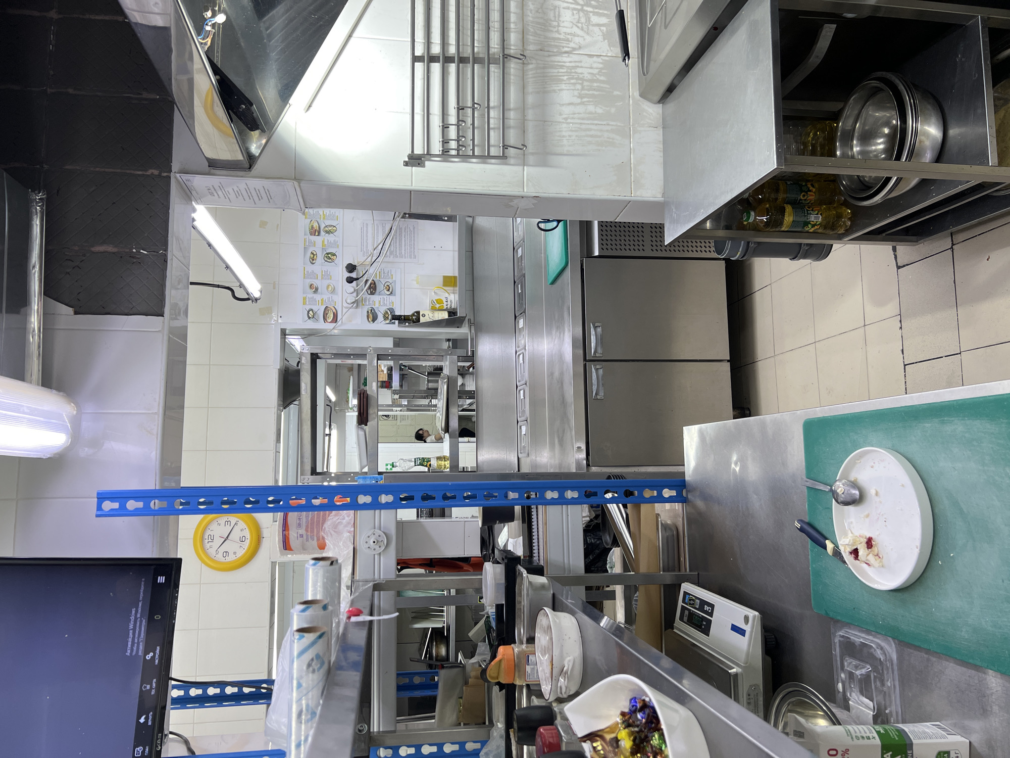 The image depicts a commercial kitchen, likely in Plastunivska, Russia, showcasing stainless steel surfaces, cooking equipment, and food preparation areas. A yellow analog clock with black numbers is visible on a tiled wall, indicating a time of approximately 12:05. On a shelf in the foreground, a white oval plate with remnants of food, a spoon, and a knife are present, resting on a green cutting board. Several bottles of cooking oil are stored on a metal shelf unit in the lower right corner. In the background, a person is partially visible in a food preparation area, and industrial shelving units are laden with various kitchen supplies. The overall environment appears to be a functional, albeit potentially messy, professional kitchen.