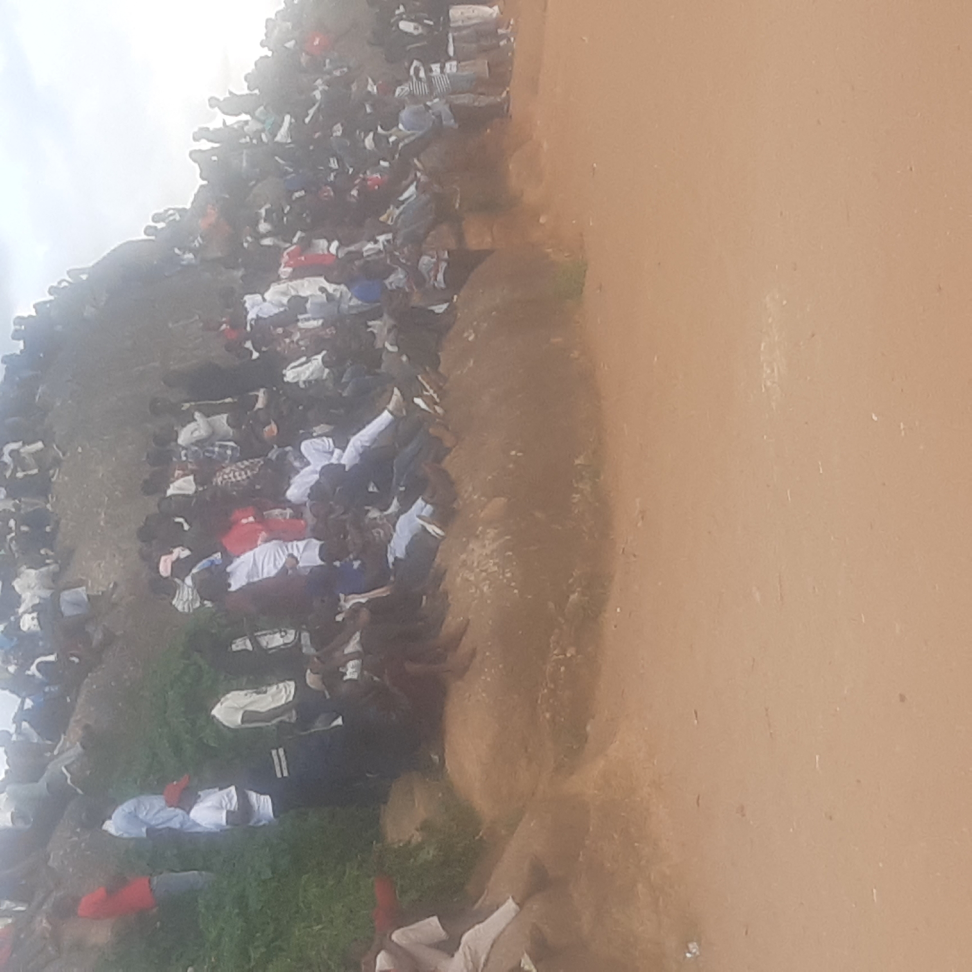 A large crowd of people are gathered on and around a rocky outcrop. Many are seated or standing on the rocks, while others are clustered at the base. The environment appears to be outdoors, with some greenery visible on the left side of the image. The ground is a sandy or dusty color. The sky is overcast. The overall impression is of a public gathering or event.