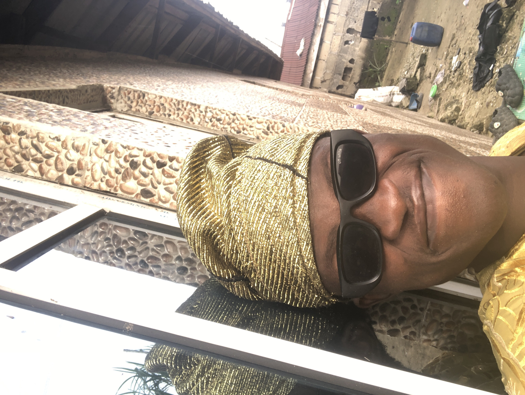 A selfie is taken outdoors in Ado, Nigeria, from a low angle. The subject is a dark-skinned person wearing a gold-colored, textured headwrap and dark sunglasses. They are wearing a gold garment. The person is positioned in front of a building with a facade made of various-sized, smooth, light-brown and gray pebbles embedded in concrete. To the left of the subject, there is a white-framed window with a reflective surface showing a partial reflection of the subject and some green foliage. To the right of the building, in the background, is a more traditional brick building with a red-tiled roof and a wooden overhang. The ground appears to be dirt and rubble, with scattered items including a blue barrel, bags of material, shoes, and discarded black plastic. The overall scene suggests a possibly rural or semi-developed urban setting.