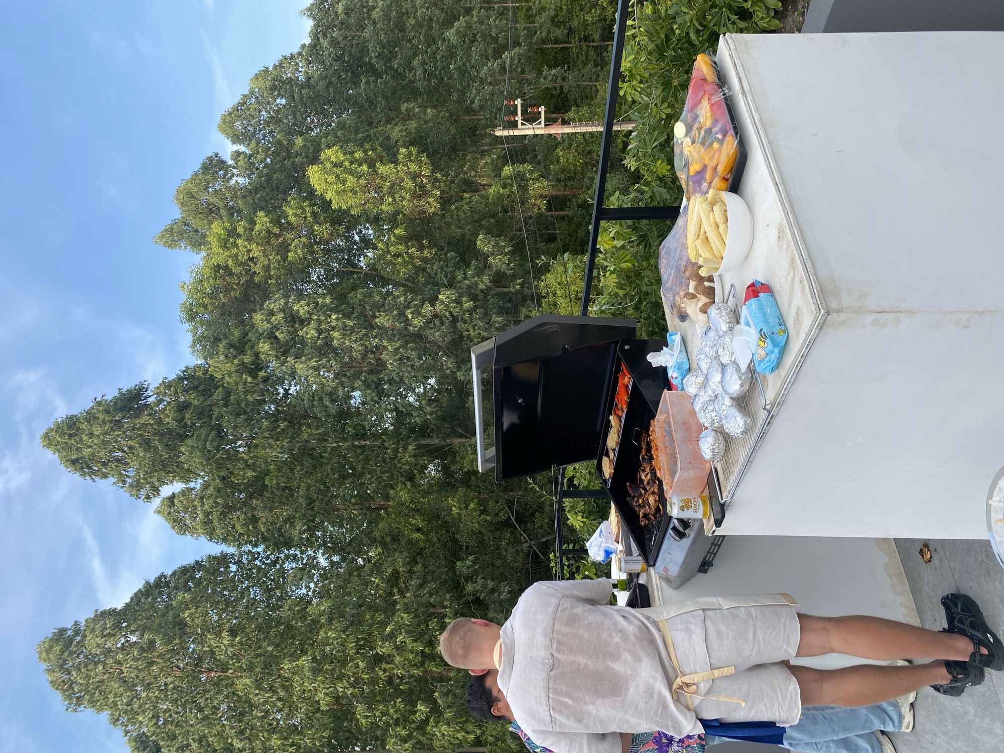 An outdoor barbecue scene unfolds under a clear blue sky, set against a dense backdrop of tall, green trees. The image captures a gathering in Choeng Thale, Thailand, during what appears to be a sunny daytime.
In the foreground, a person with short, light hair is visible from the back, wearing a light-colored shirt, shorts, and an apron, actively tending to a black barbecue grill. Another person is partially visible next to them. The grill is laden with various items, including meat skewers and chopped bell peppers, all in the process of cooking.
Adjacent to the grill, a white counter or table holds an assortment of food and provisions. These include plastic-wrapped bell peppers in red and yellow, a white bowl filled with baby corn, a cluster of mushrooms, and several items individually wrapped in aluminum foil, likely potatoes. Blue plastic bags or wrappers are also present on the table. A "SINGHA" beer can is visible near the grill.
The background is dominated by lush, green foliage, primarily tall, slender trees that reach towards the sky. A utility pole with power lines can be seen extending through the trees, hinting at the area's infrastructure. The overall atmosphere suggests a casual and relaxed outdoor cooking event. Small text, possibly "80GM," is discernible on one of the blue plastic bags on the table.