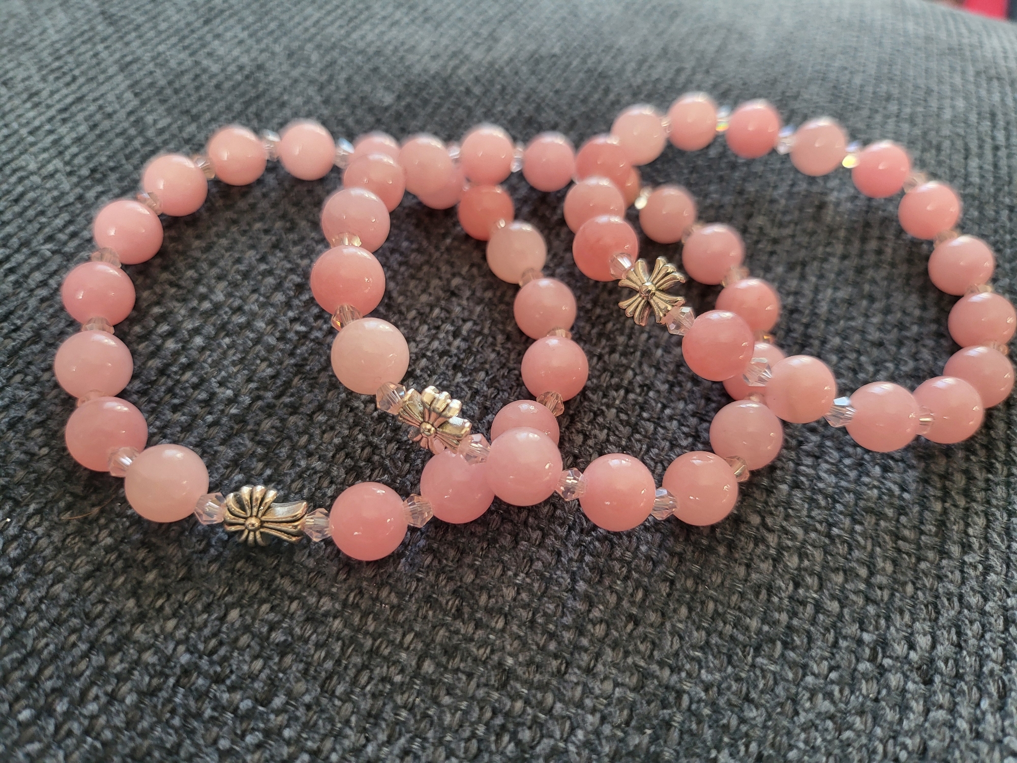 Three pink beaded bracelets are laid out on a textured gray surface. Each bracelet is composed of round, opaque pink beads, interspersed with smaller, clear faceted beads. Three metallic charms, shaped like stylized flowers or stars, are present, one on each bracelet. The bracelets are positioned in a loosely overlapping arrangement. The background is a dark gray fabric with a visible woven texture. The lighting is natural, casting soft shadows. The image does not contain any information related to the city of Peski, Russia.