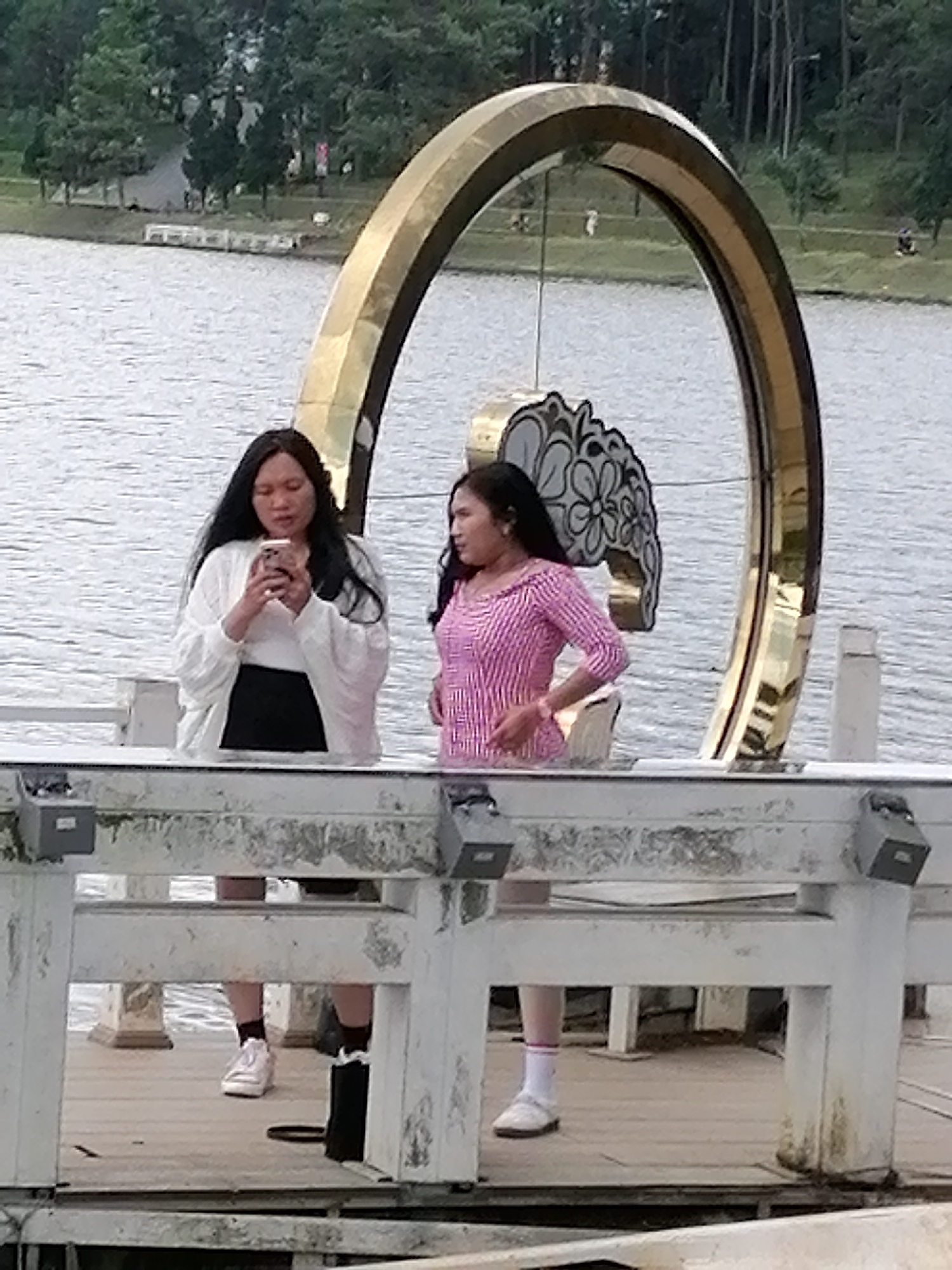 The image shows two women standing on a wooden pier near Xuan Huong Lake in Da Lat, Vietnam. A large, gold, arch-shaped sculpture is visible behind them, along with a floral decoration. The woman on the left is wearing a white top and a black skirt, holding a smartphone. The woman on the right is wearing a pink striped shirt. The lake is in the background, with trees and foliage on the far shore. The pier has white railings and wooden planks. The overall lighting suggests an overcast day.