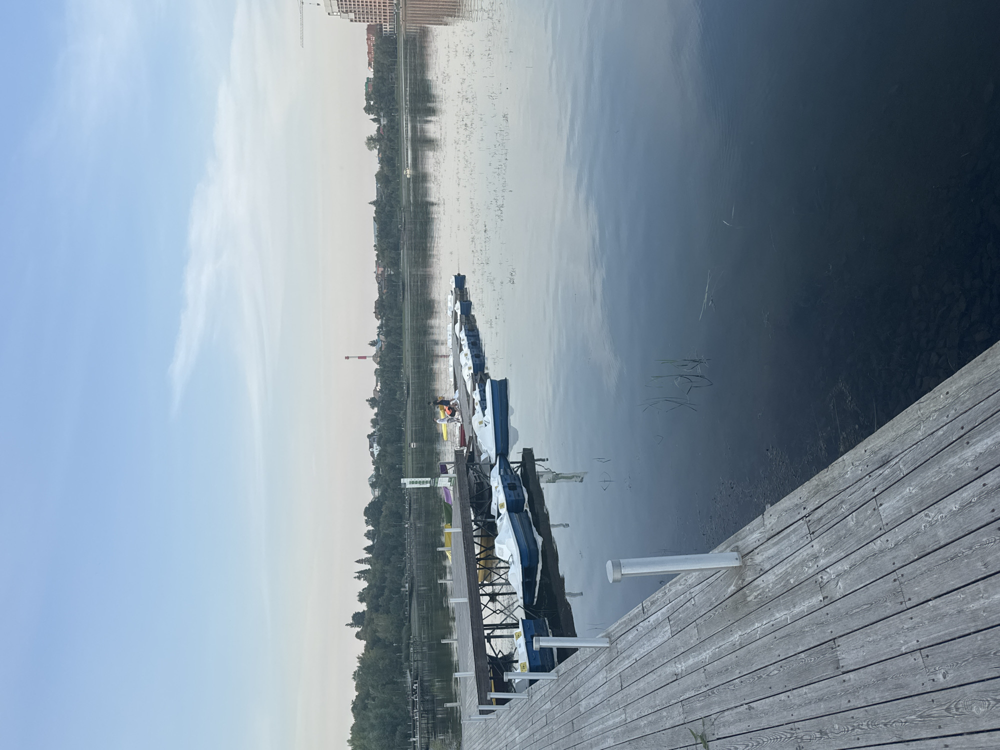 The image depicts a serene lake scene at the Ufa city embankment, Russia. A wooden pier juts out into the calm water, where several small pedal boats are moored. The boats, white with blue accents, are neatly lined up, suggesting a rental facility. In the background, a lush green treeline borders the opposite bank, dotted with residential buildings and industrial structures, including a prominent red and white striped smokestack. The sky is a pale blue, with thin wisps of clouds, indicating either early morning or late evening. Reflections of the sky and the boats are visible on the water's surface. Two individuals are visible walking on the pier, appearing relaxed and enjoying the tranquil atmosphere. The overall impression is one of a peaceful recreational area within an urban setting.