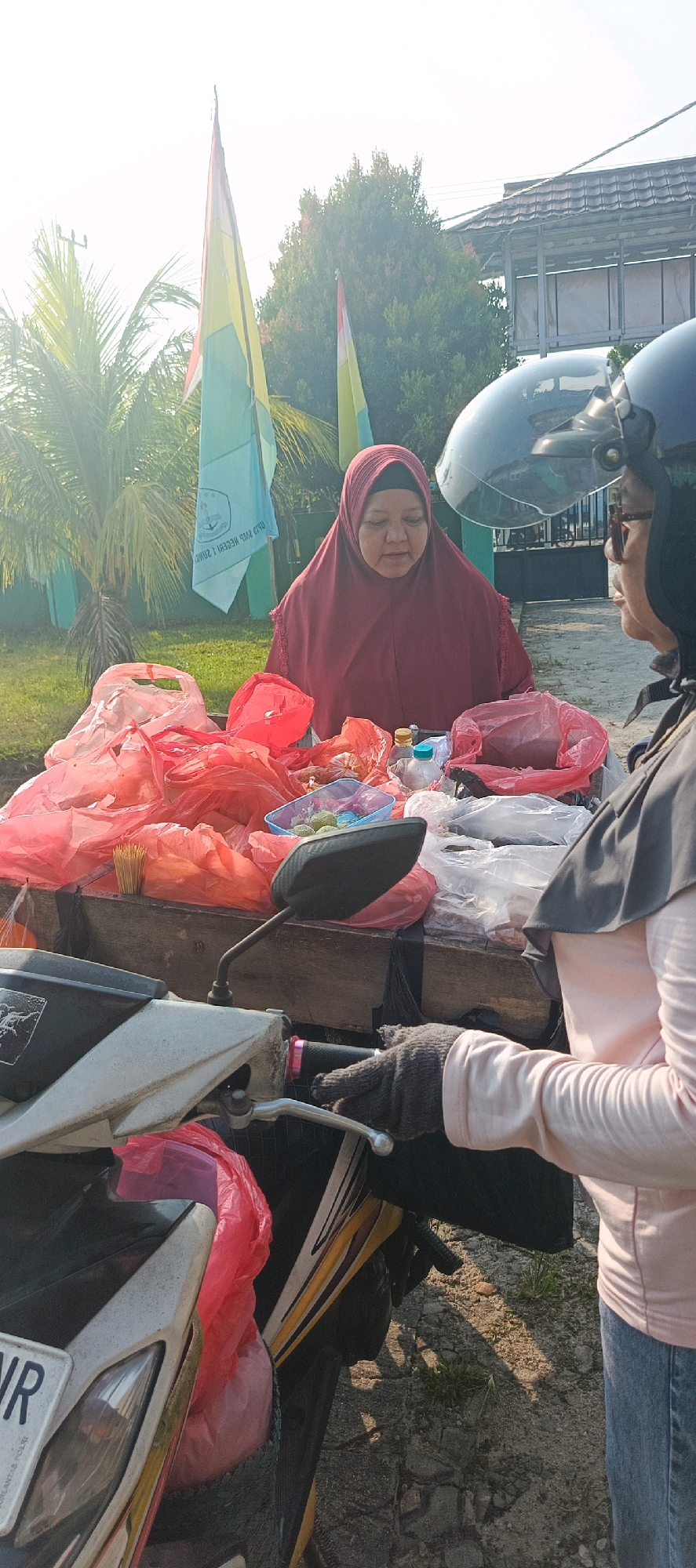 In the city of Desa Sarang Mandi, Indonesia, a vendor wearing a red hijab and a black headscarf stands behind a wooden cart filled with various items, predominantly red plastic bags containing food or goods. She is looking down at the items on the cart. To her right, a second individual, also wearing a hijab and a dark head covering, is positioned in front of the cart, partially obscured by a motorcycle. This second person is wearing a light pink long-sleeved shirt and jeans, and a dark grey glove on their left hand, which is resting on the motorcycle's handlebar. A motorcycle, with a portion of its front visible and a license plate, is parked in the foreground. Behind the cart, two flags are visible, one light blue and one yellow, with trees and a building in the background. The scene is outdoors, likely during daylight, with sunlight casting shadows.