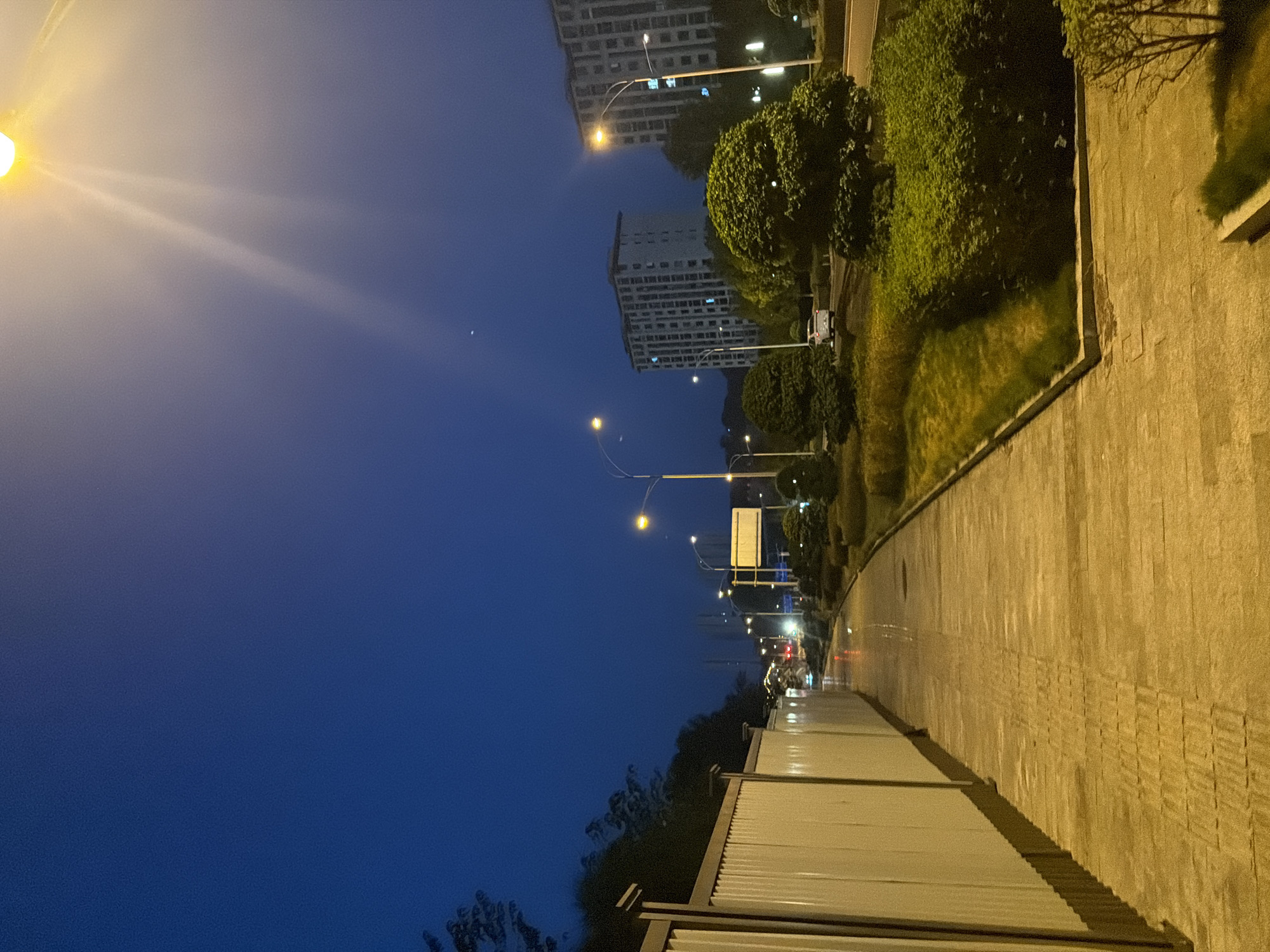 This is a nighttime photograph of an urban scene in Jiangjin, China. The sky is a deep blue, transitioning to a lighter hue near a bright streetlight in the upper right corner, which casts a strong glow and visible light rays. The foreground features a paved pedestrian walkway with a textured surface, possibly illuminated by artificial lights that create a yellowish cast. To the left of the walkway is a tall, white corrugated metal fence. Beyond the fence, trees are visible against the dark sky.
Further into the scene, the walkway curves and leads towards a street with streetlights illuminating the path. There are several streetlights along the road, some with orange-yellow illumination. To the right of the walkway, a grassy area and manicured bushes are present, bordered by a raised concrete edge. In the middle ground, a white car is visible on the road. Across the road, there are multi-story buildings with illuminated windows, suggesting residential or office spaces. The overall environment suggests a well-maintained urban area, possibly a park or a residential complex, under the twilight or early night sky.