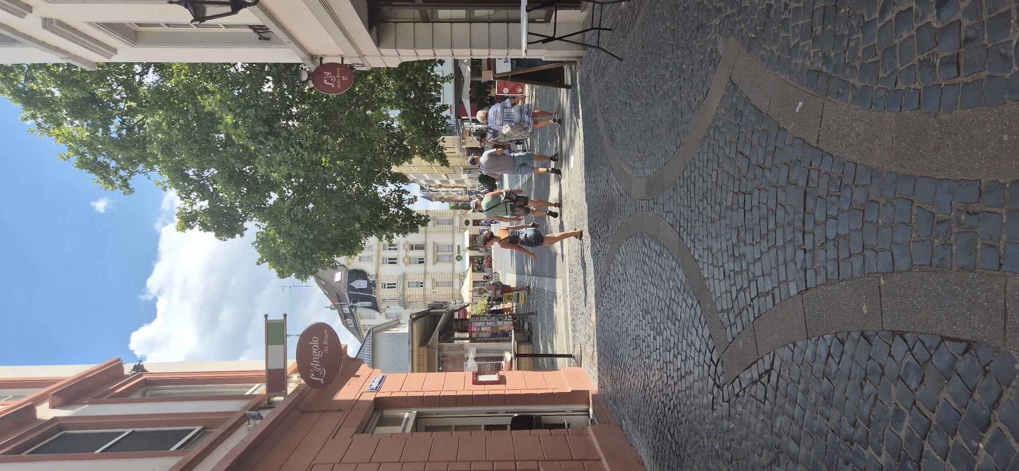 This is a ground-level, eye-level, vertical, full shot of a pedestrian street scene in Mainz, Germany, on a sunny day with some clouds. The street is paved with dark cobblestones, with curved inlays of lighter material. On the left is a reddish-brown building with white trim. A sign with an Italian flag motif hangs above the entrance to "L'Angolo du Biagio". Across the street, several people are walking away from the camera down the street. The group includes a woman in a blue and white patterned dress, a man in a grey t-shirt and blue shorts, a man in a green t-shirt and patterned shorts, and a woman in a denim skirt. Further down the street, more people, shops, and buildings are visible. A large green tree is prominent in the middle ground.