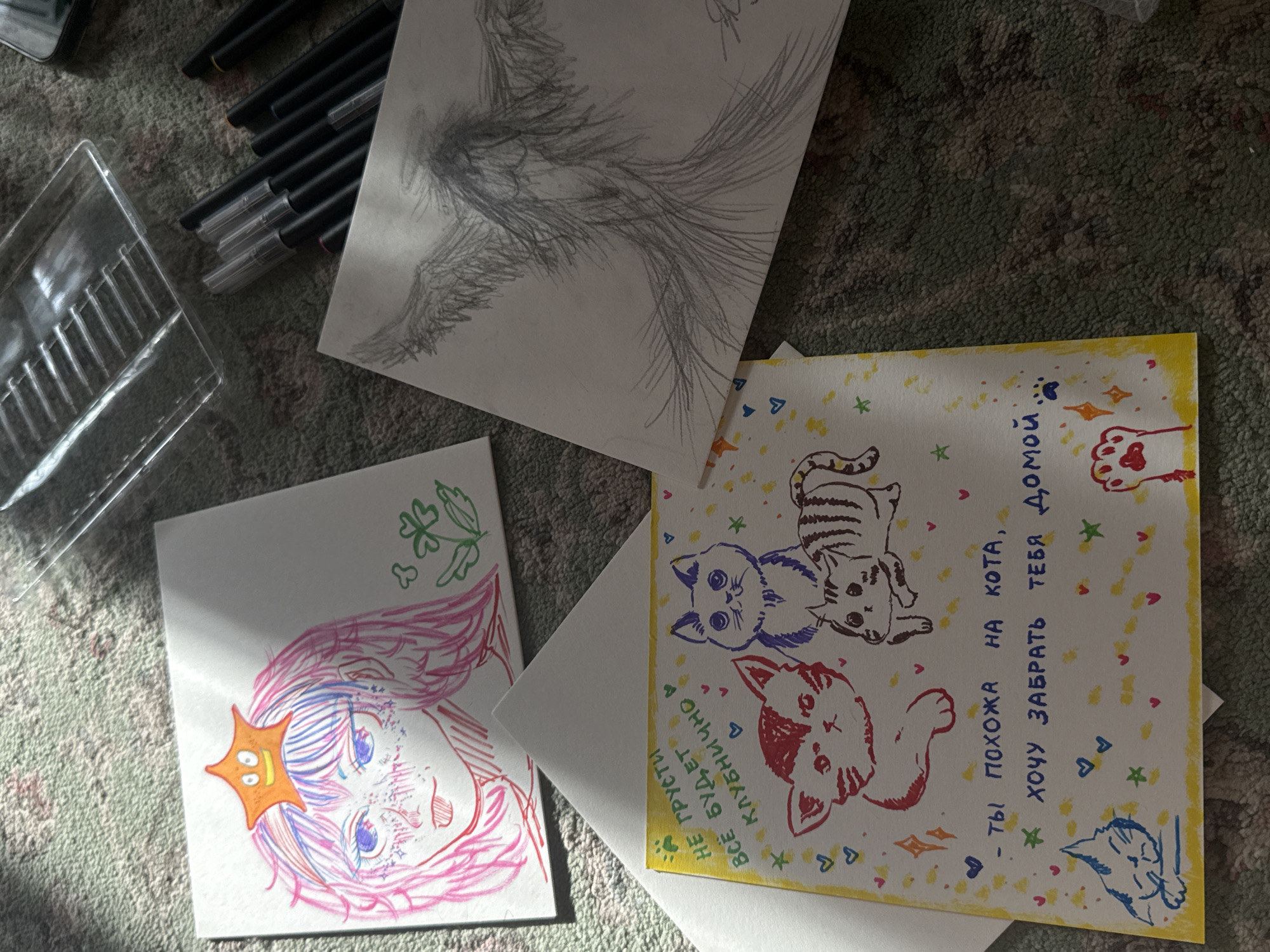 The image shows a collection of drawings and art supplies scattered on what appears to be a textured surface, possibly a rug.
There are four main paper-based artworks:
1. A drawing of wings in pencil.
2. A colorful drawing of a female character with pink hair, blue eyes, and a star on her head.
3. A postcard-style drawing with multiple cats in different colors and patterns. The background is decorated with stars, hearts, and other small motifs. There is text in Russian on the postcard.
4. A blank sheet of paper.
Art supplies are visible: a plastic container and a collection of black pens or markers.
The setting appears to be indoors. There's no indication of a specific location or connection to Uryupinsk, Russia.