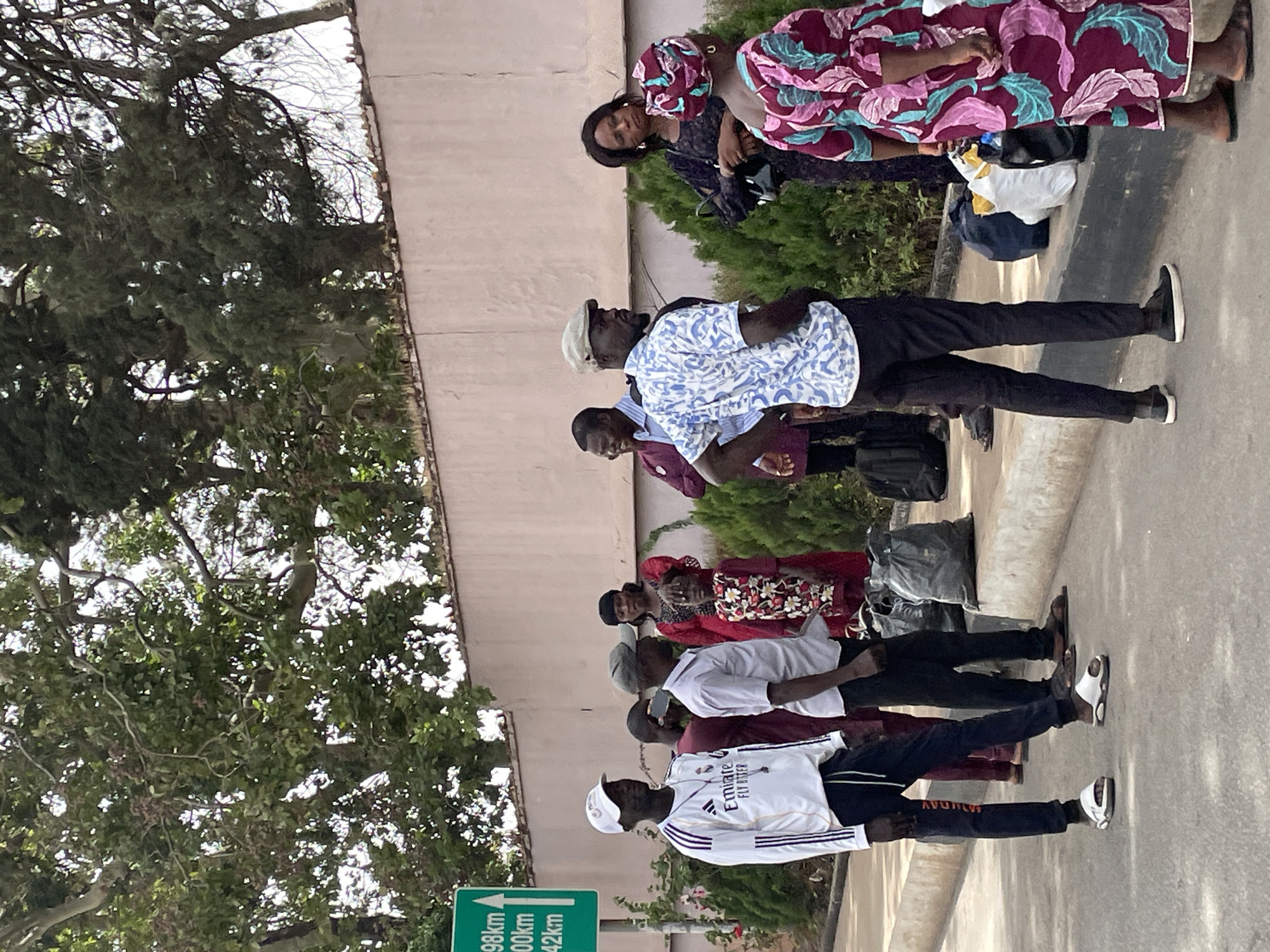 A line of people stands on a road next to a curb. A sign indicates locations are 98 km, 100 km, and 142 km away, suggesting they may be waiting for transportation. The people are of African descent, and their clothing includes both modern and traditional styles. The location is likely Jos, Nigeria, given the clothing styles and context. The setting appears to be outdoors, with trees and vegetation visible.