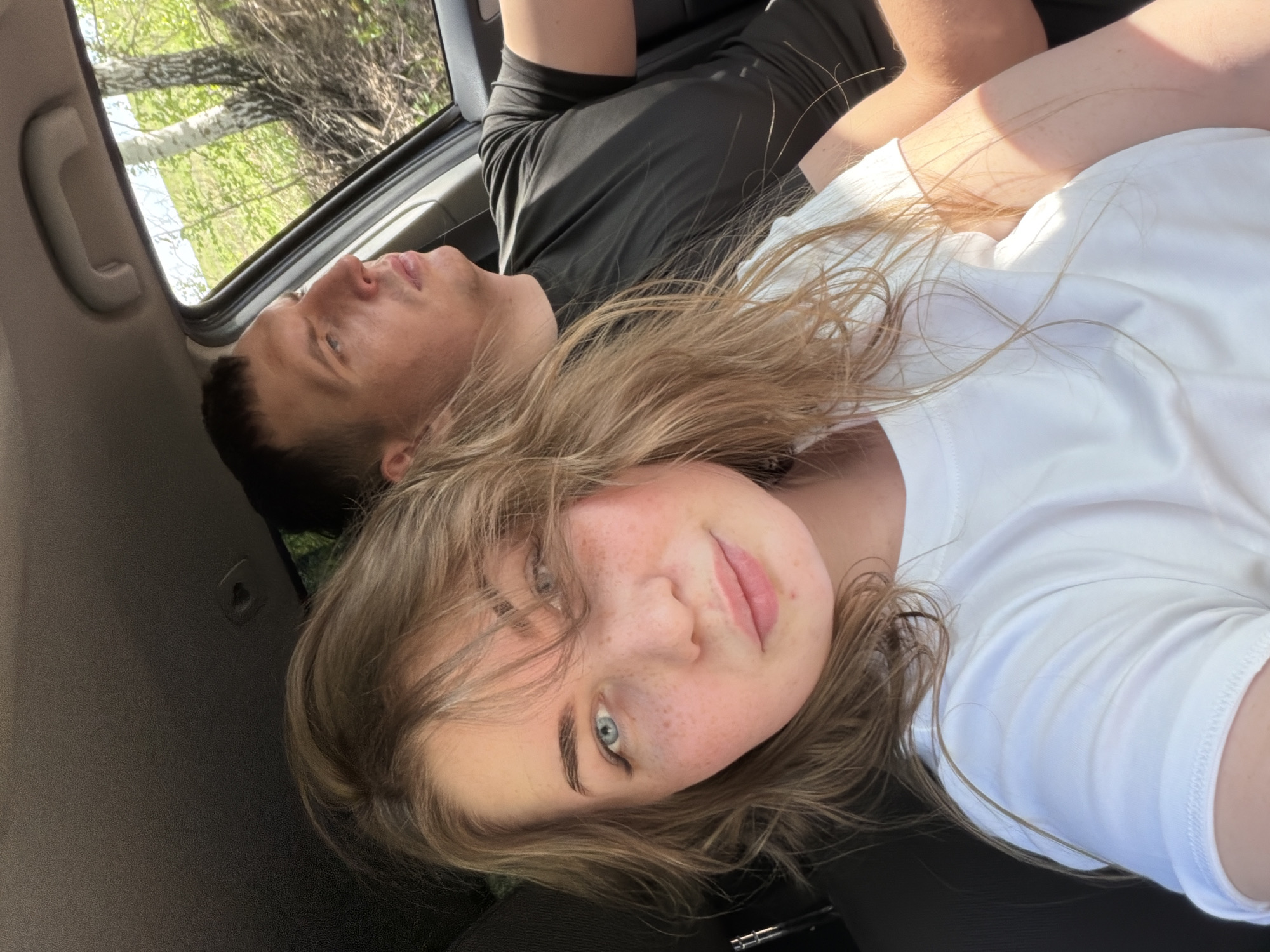 A selfie was taken inside a vehicle, likely a car, with two people visible. A fair-skinned young woman with freckles, light brown hair, and blue eyes is the primary subject. She is wearing a white t-shirt and is positioned closer to the camera. Behind her, a fair-skinned young man with short dark hair is lying down. He is wearing a dark-colored t-shirt and is looking out the sunroof. Trees are visible through the sunroof. The location of the scene is in the city of Ural'skoe, Russia.