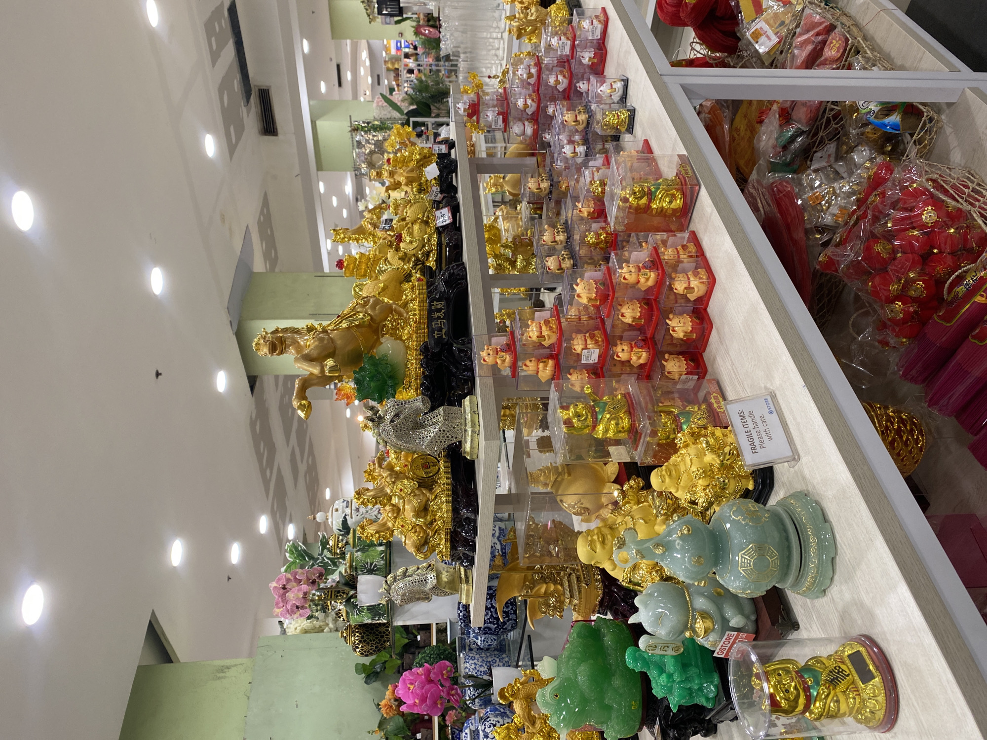 The image depicts a vibrant retail display of numerous auspicious figurines and decorative items within a Gaisano Grand Mall in Davao City, Philippines. The scene is brightly lit by recessed circular lights in a white ceiling, with light green structural columns or walls visible in the background, characteristic of a large department store.
The central focus is a multi-tiered display counter laden with golden and jade-green statues and figurines. The top shelf features large, ornate golden statues, including a prominent arrangement of eight golden horses symbolizing prosperity and a silver-colored horse statue. Alongside these are large, sculpted jade-green figures, likely money toads or Buddha statues. Artificial pink orchids and other floral arrangements, along with decorative ceramic vases in blue and white patterns and black and green geometric patterns, adorn the background of this shelf.
The middle shelf is predominantly filled with arrays of smaller, golden Maneki-neko (waving lucky cat) figurines, each individually packaged in clear plastic boxes with red bases, stacked in organized pyramids. Larger golden laughing Buddha figures and other golden animal statues are also visible on this shelf, some with visible price tags.
The bottom shelf showcases various medium-sized jade-green and golden figurines, including several laughing Buddhas, a light blue/grey cat-like figure holding a gold coin, and a decorative light green gourd-shaped object. A smaller golden Maneki-neko, housed in a clear cylindrical container, is positioned on the far left. A white sign with black text reading "FRAGILE ITEMS: Please handle with care." and displaying a "GSTORE" logo is clearly visible on this shelf.
Below the bottom shelf, several woven baskets are filled with packaged red and gold-colored items, appearing to be festive decorations like lanterns and other celebratory goods. No people are visible in the immediate foreground of the image.
