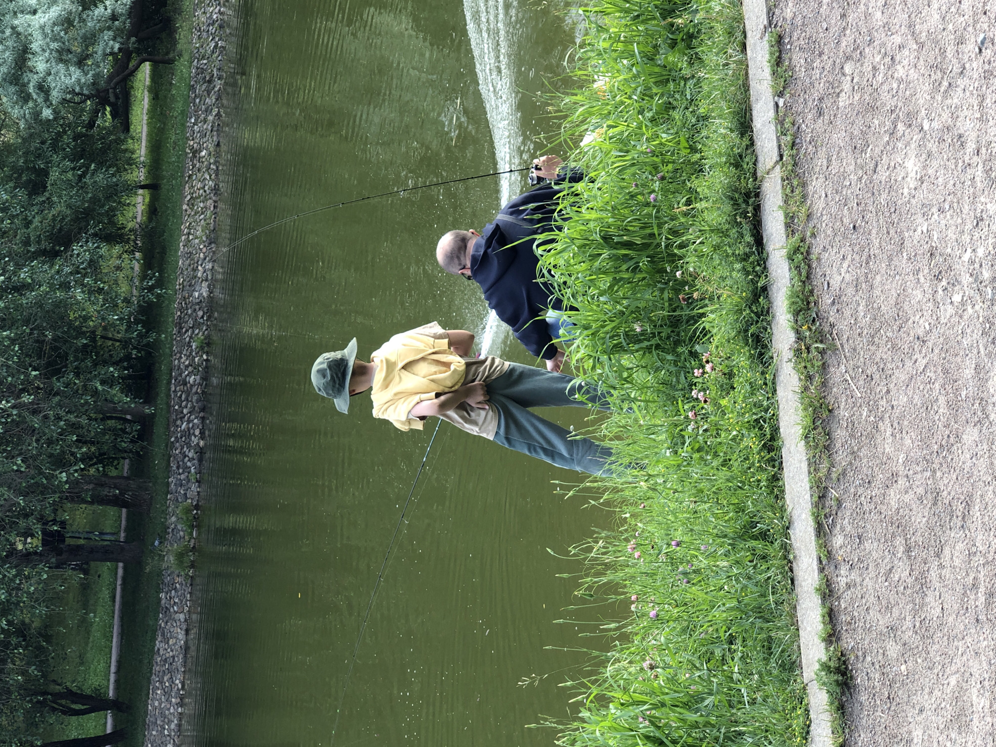 Two people are fishing by a narrow, calm body of water. The person closer to the viewer is wearing a yellow shirt and a green bucket hat. They are holding a fishing rod and looking out at the water. The second person, farther away, is wearing a dark blue hooded jacket and jeans. They are also holding a fishing rod, casting into the water. The water has a slightly green hue and appears calm, with ripples visible in some areas. The bank of the water is lined with lush green grass and some small pink flowers. A stone embankment separates the water from a paved walkway on the right side of the image. Trees are visible on the opposite bank of the water, suggesting a park or natural setting. The lighting suggests it is daytime, likely overcast or late afternoon, as there are no strong shadows. The overall scene conveys a peaceful and relaxed atmosphere.