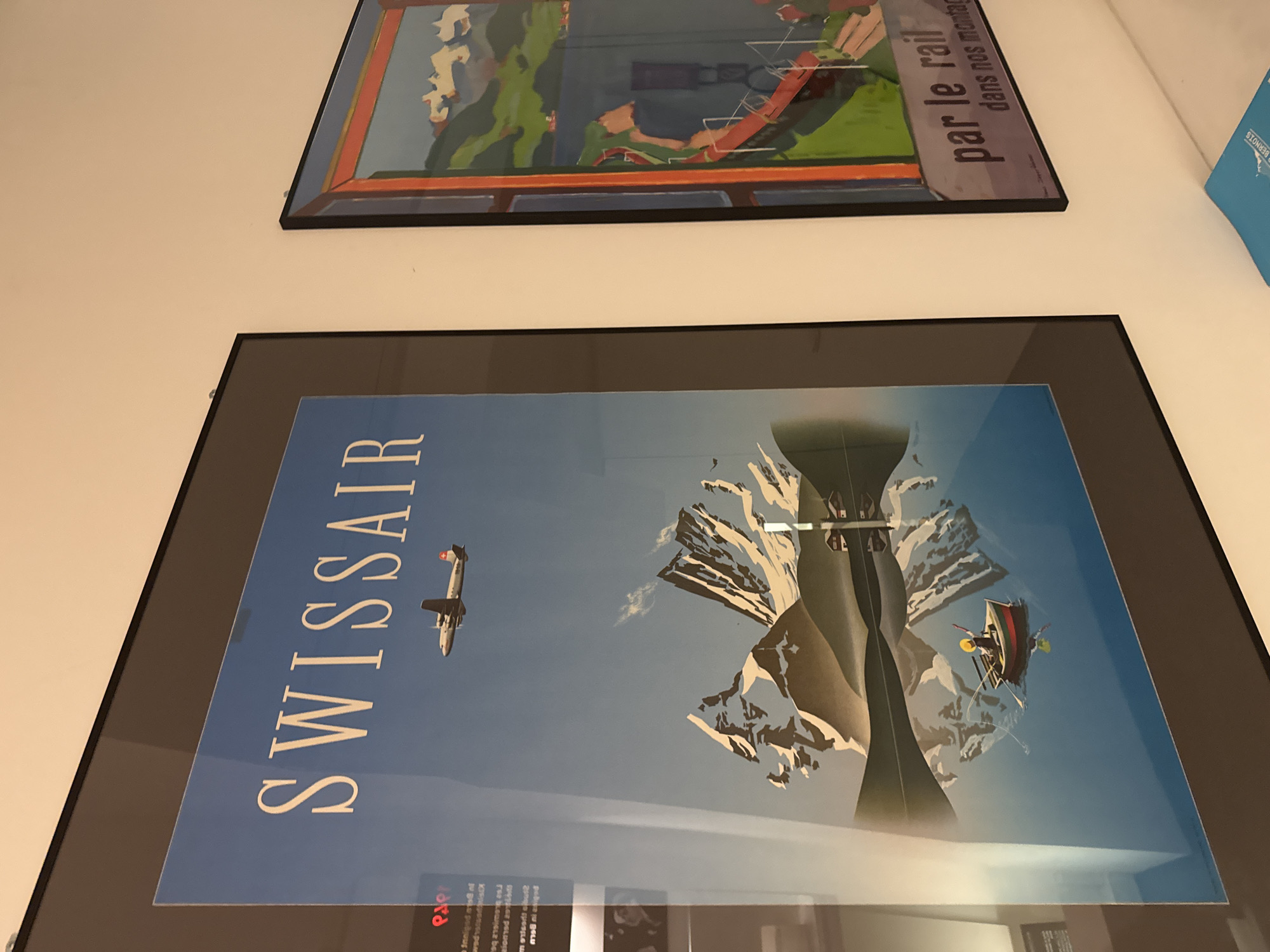 The image shows two framed posters hanging on a white wall. The poster on the left features a blue sky with a white airplane flying above snow-capped mountains reflected in a body of water. A small boat with a single person fishing is visible in the water. The word "SWISSAIR" is prominently displayed at the top of the poster in white lettering. The poster on the right has a colorful illustration of a mountainous landscape with a red bridge and a body of water. Text at the bottom of this poster reads "par le rail dans nos montag". The overall scene appears to be an interior wall, likely in a gallery or a home.