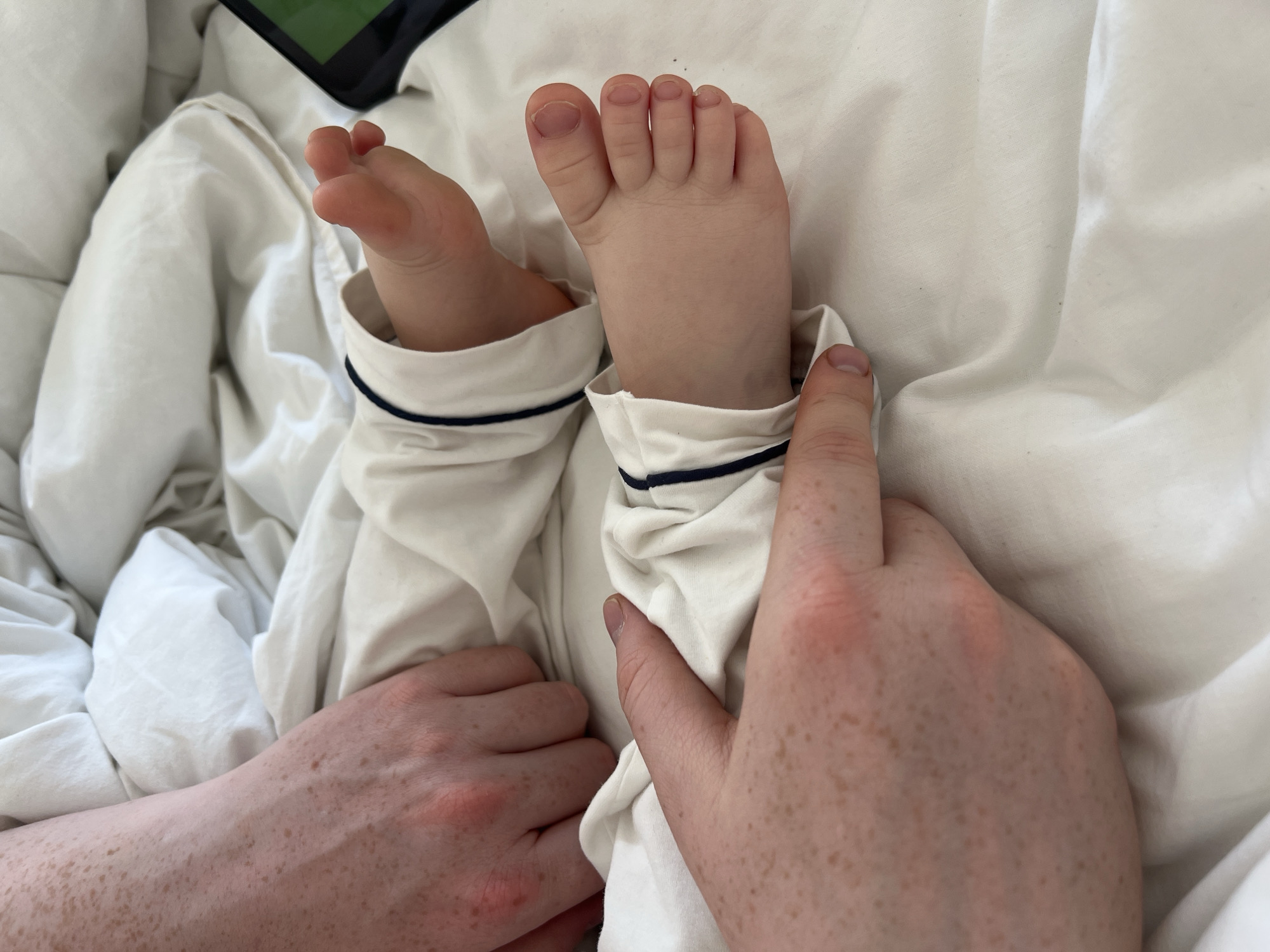 The image depicts two baby feet clad in white pajamas with navy trim, lying on a white bed sheet. An adult hand with freckles is holding one of the baby's ankles. The photo is taken from an overhead perspective. The location is unknown, there is no evidence of the city Prudki, Russia.