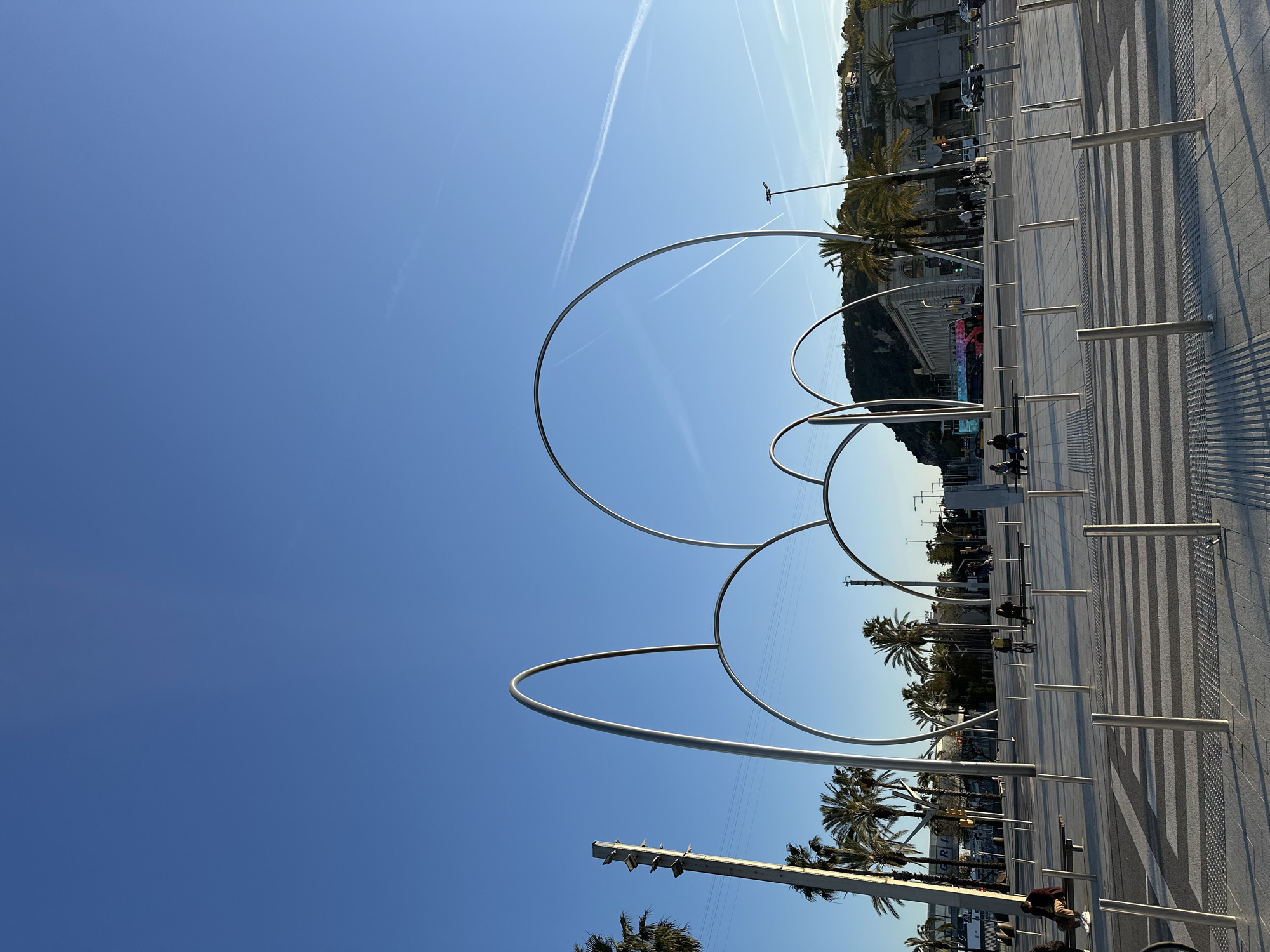 The image captures a street scene in Barcelona, Spain, featuring distinctive circular metal sculptures along the promenade. The sculptures, arranged in an overlapping sequence, stand out against the clear blue sky. The promenade itself is characterized by a striped pavement design and metal bollards. Palm trees line the street, adding to the city's characteristic landscape. Buildings are visible in the background, suggesting an urban environment. Pedestrians are present along the street, some walking and others seated on benches.