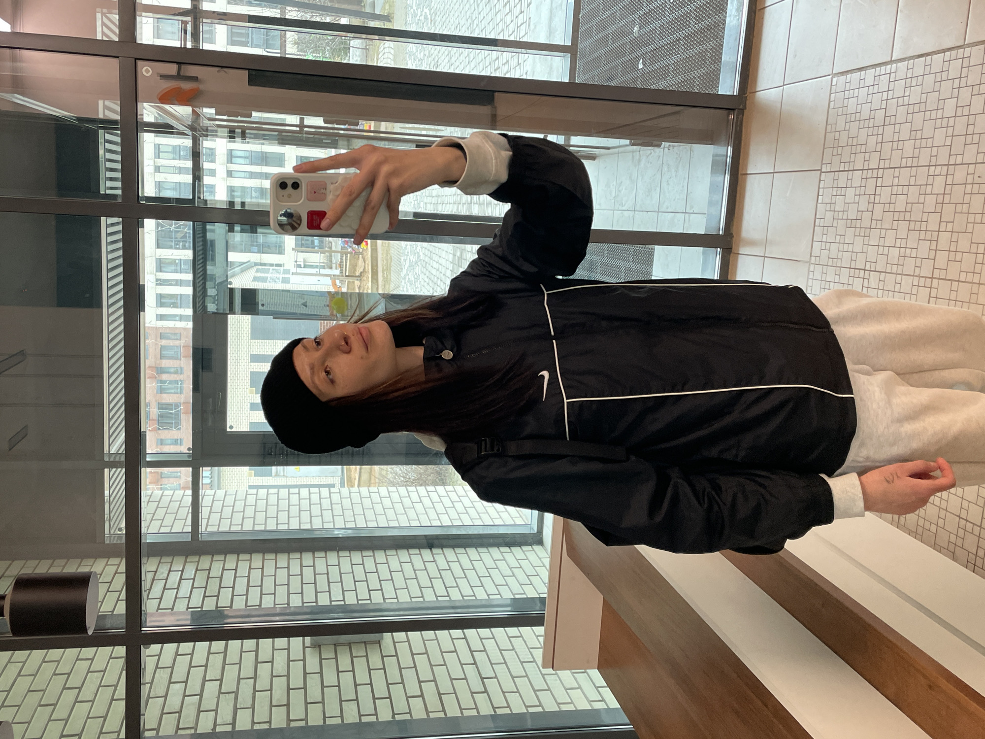 A woman is taking a selfie in what appears to be the entrance area of a building. She is wearing a black jacket with a white Nike logo, light-colored sweatpants, and a black beanie. She is holding a white smartphone with stickers on the back. The background consists of glass windows and doors, revealing exterior views of buildings and the street. The floor and walls are tiled. It's not possible to verify the exact location in Izhevsk, Russia, from the image alone.