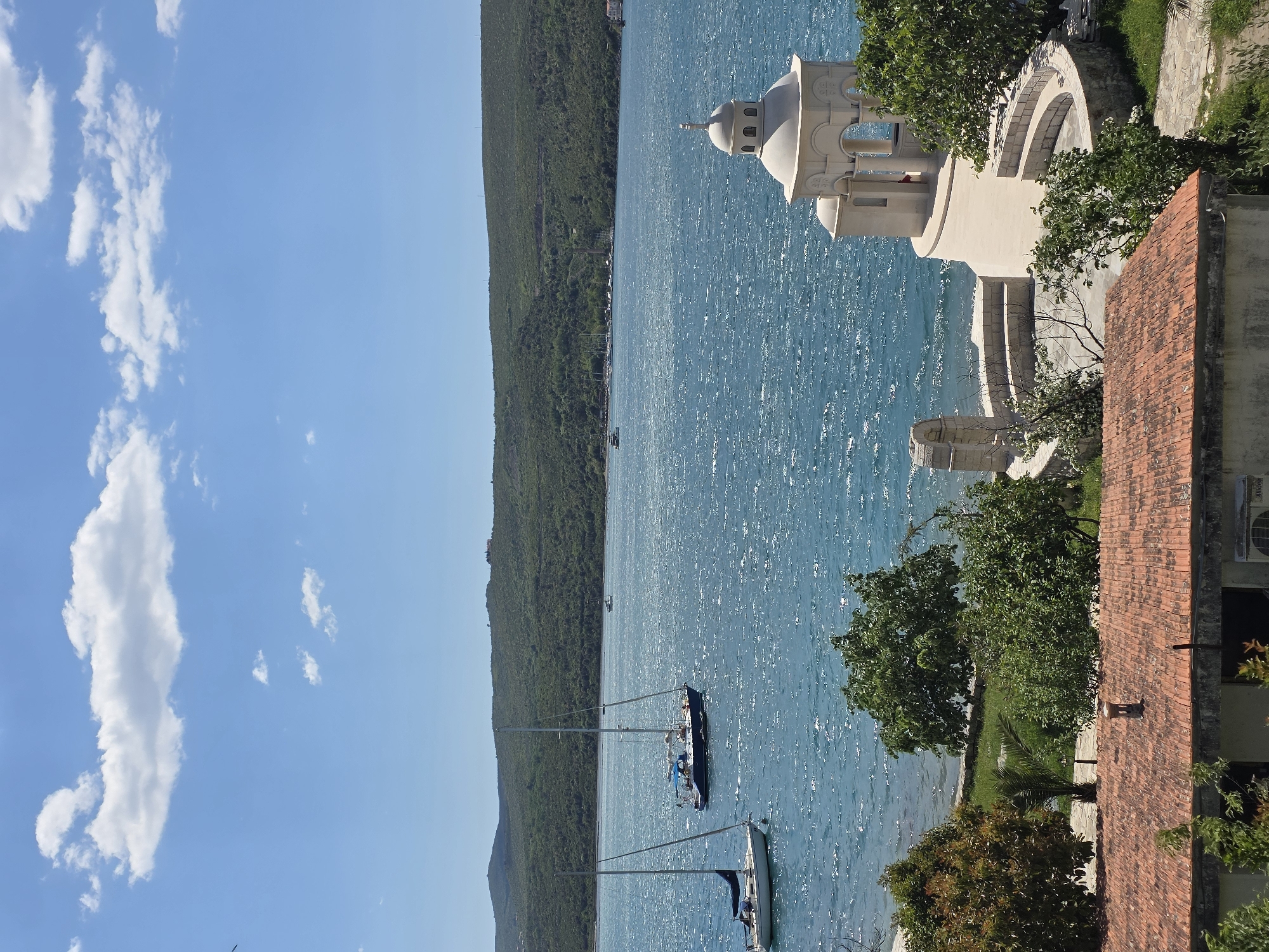The image shows a view of Đuraševići, Montenegro. The blue water of the bay is visible, with two sailboats anchored near the shore. A green, mountainous coastline stretches along the left side of the image. On the right, there is a cream-colored stone structure, possibly part of a church or other religious building, with distinctive domed towers and arched openings. Nearby, there is a terracotta-tiled roof of a building with trees scattered around. The sky is blue with some scattered white clouds.