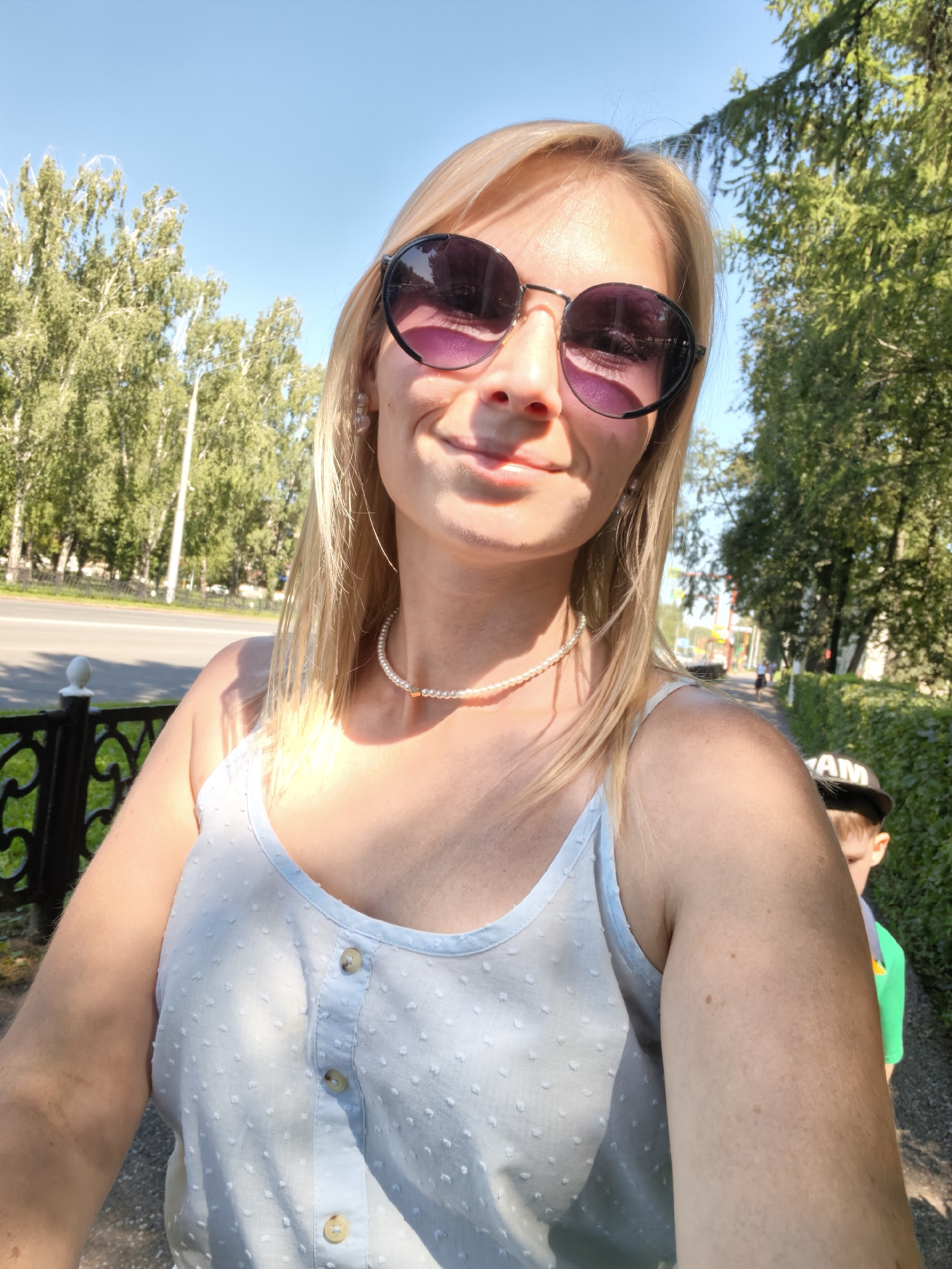 A woman with blonde hair and sunglasses is taking a selfie outdoors on a sunny day. She is wearing a light blue, button-up tank top and a pearl necklace. Behind her, a street with trees lining the sidewalk is visible. A child in a green shirt and a black baseball cap is partially visible to her right. The location is the city of Sterlitamak, Russia.