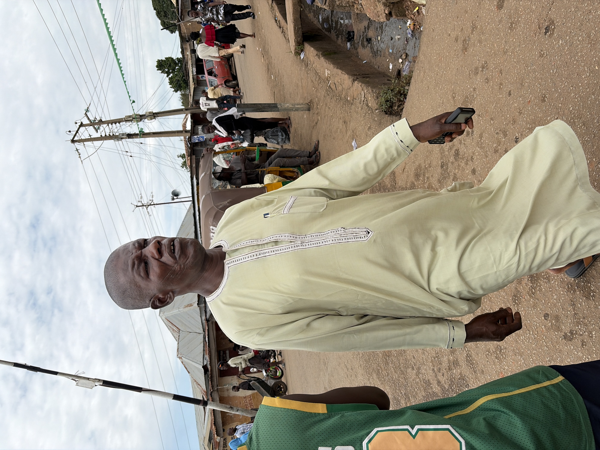 The image depicts a man in light green traditional attire walking on a dirt road in Gura Top, Nigeria. He is bald and has a prominent scar on his face. He is holding a black object, possibly a phone, in his right hand. The man is walking towards the right side of the frame. In the background, other people, vehicles, and buildings are visible, suggesting a busy street scene. There is a drainage ditch on the right side of the road filled with debris. Electrical poles and wires are prominent in the sky. The overall lighting suggests it is daytime with overcast skies.