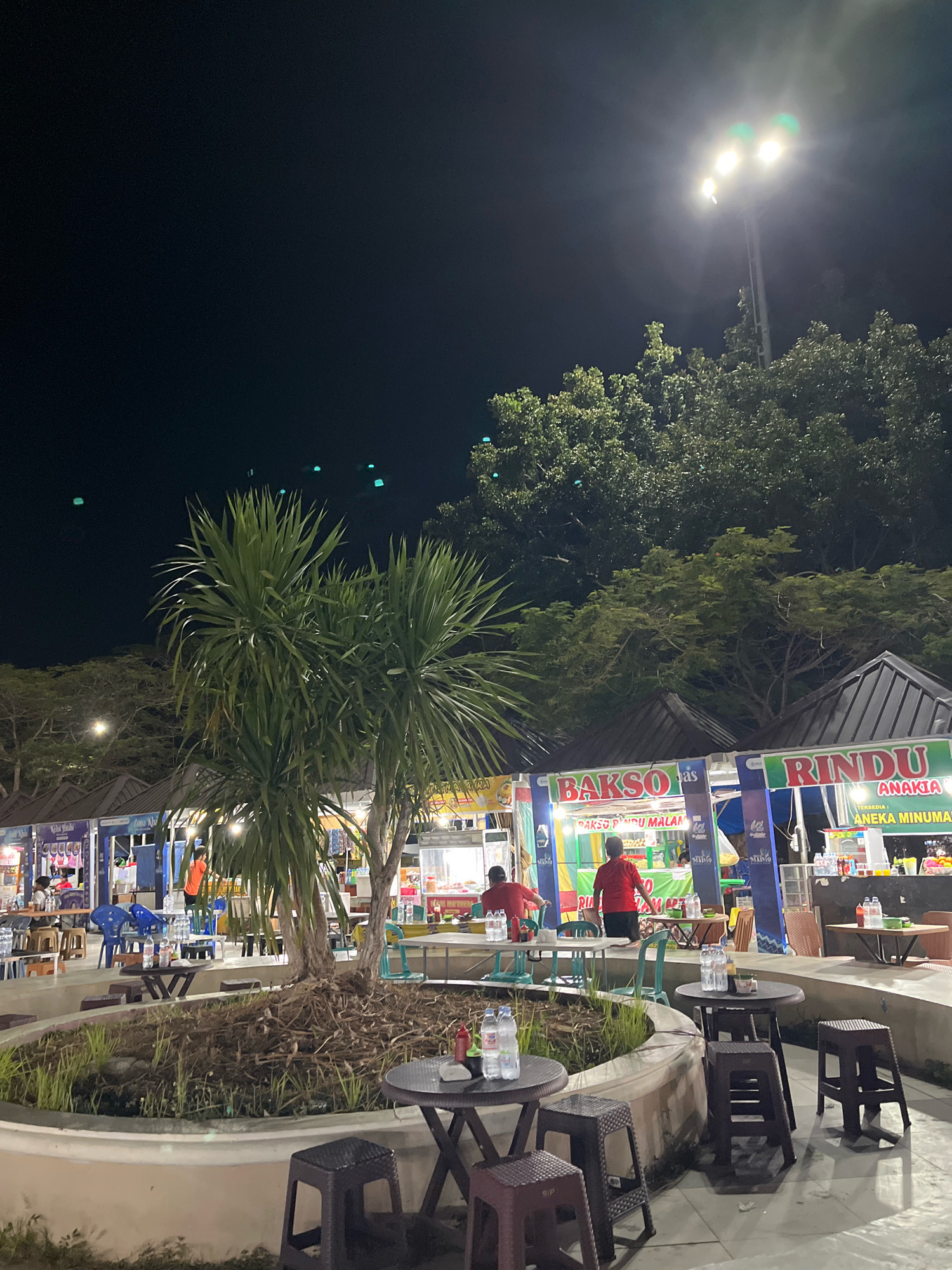 The image depicts a nighttime scene at a food court in Kendari, Indonesia. The food court features several stalls with illuminated signs advertising food and drinks. In the foreground, there are several round tables with chairs, some set up for dining with bottles of water and condiments. A planter with greenery and dry vegetation is situated in the center. In the background, people are seen at the food stalls, some appearing to be staff preparing food and others as customers. The overall atmosphere is that of a casual outdoor dining area, typical of evening street food establishments in the city.