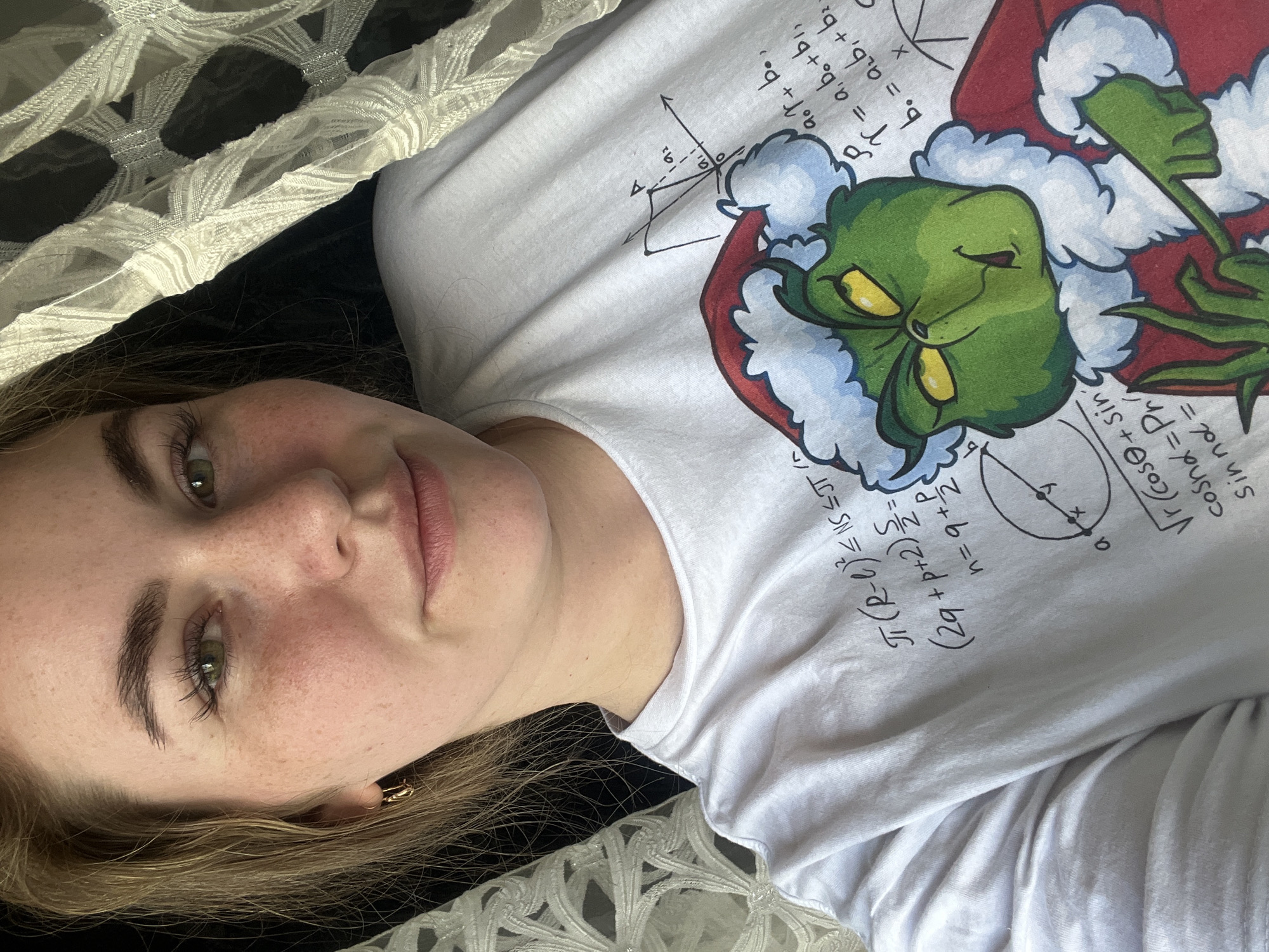 A fair-skinned person with light brown hair and green eyes is lying on a white crocheted hammock or bed covering with black background. The person is wearing a white long-sleeved shirt with an image of the Grinch and mathematical formulas on it. The image is cropped close to the person's face and upper body. No location information is available.