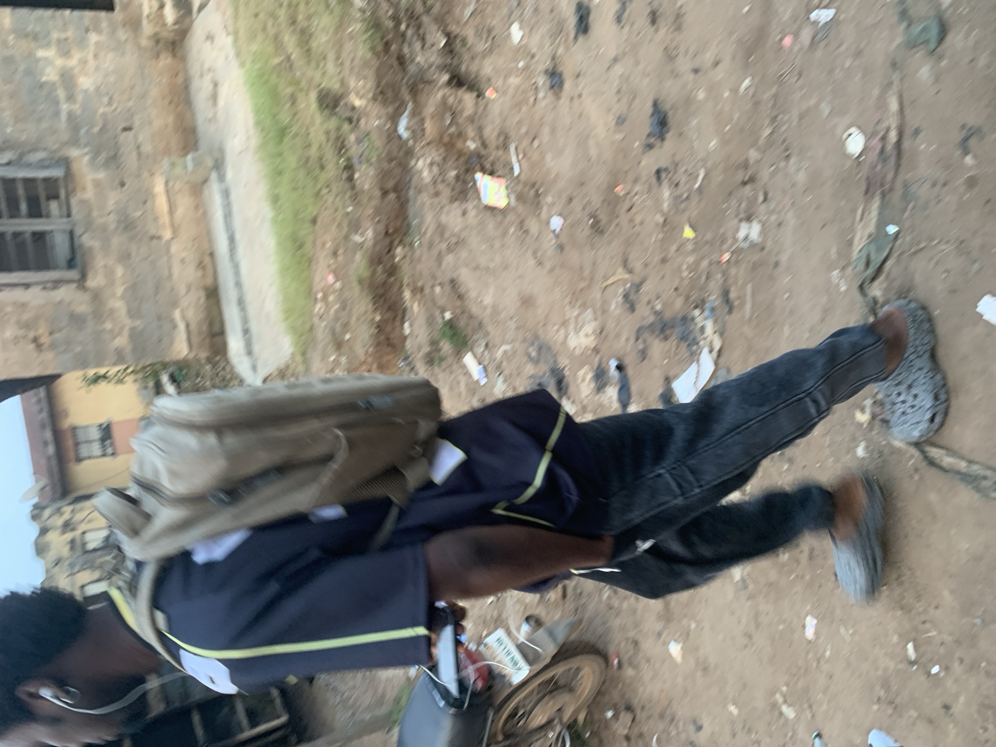 A person walks down a dirt road in Shagamu, Nigeria, wearing a dark blue t-shirt with light green stripes, dark denim jeans, and grey Crocs. They carry a tan backpack and have white earbuds in their ears. A motorcycle is parked to the left of the frame, and a building with a yellow facade and barred windows is visible in the background. The ground is littered with debris.