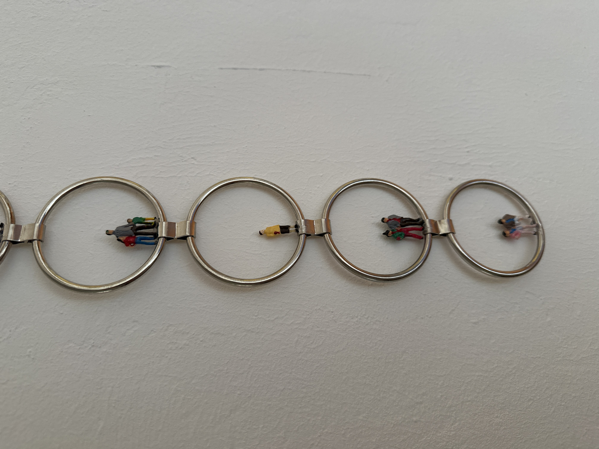 The image depicts a close-up, eye-level, vertical shot of a chain of four interconnected metal rings mounted on a textured, off-white wall. Each ring contains a group of miniature figures, arranged as if interacting or standing together.
Starting from the top ring, a family of three is visible: a man in a grey jacket, a woman in a red jacket, and a child in a green shirt.
The second ring from the top features a single miniature figure wearing a yellow jacket and black pants.
The third ring from the top shows another group of three figures, similar in composition to the top ring, with one in a red jacket, one in a green jacket, and one in a grey jacket.
The bottom ring contains a group of three miniature figures: one in a blue jacket, one in a pink jacket, and one in a dark blue top.
The metal rings are a dull silver color and appear to be attached to each other via a connecting metal piece. The wall's texture is rough and uneven. No specific location in Ammeldingen bei Neuerburg, Germany, is identifiable from the image itself.