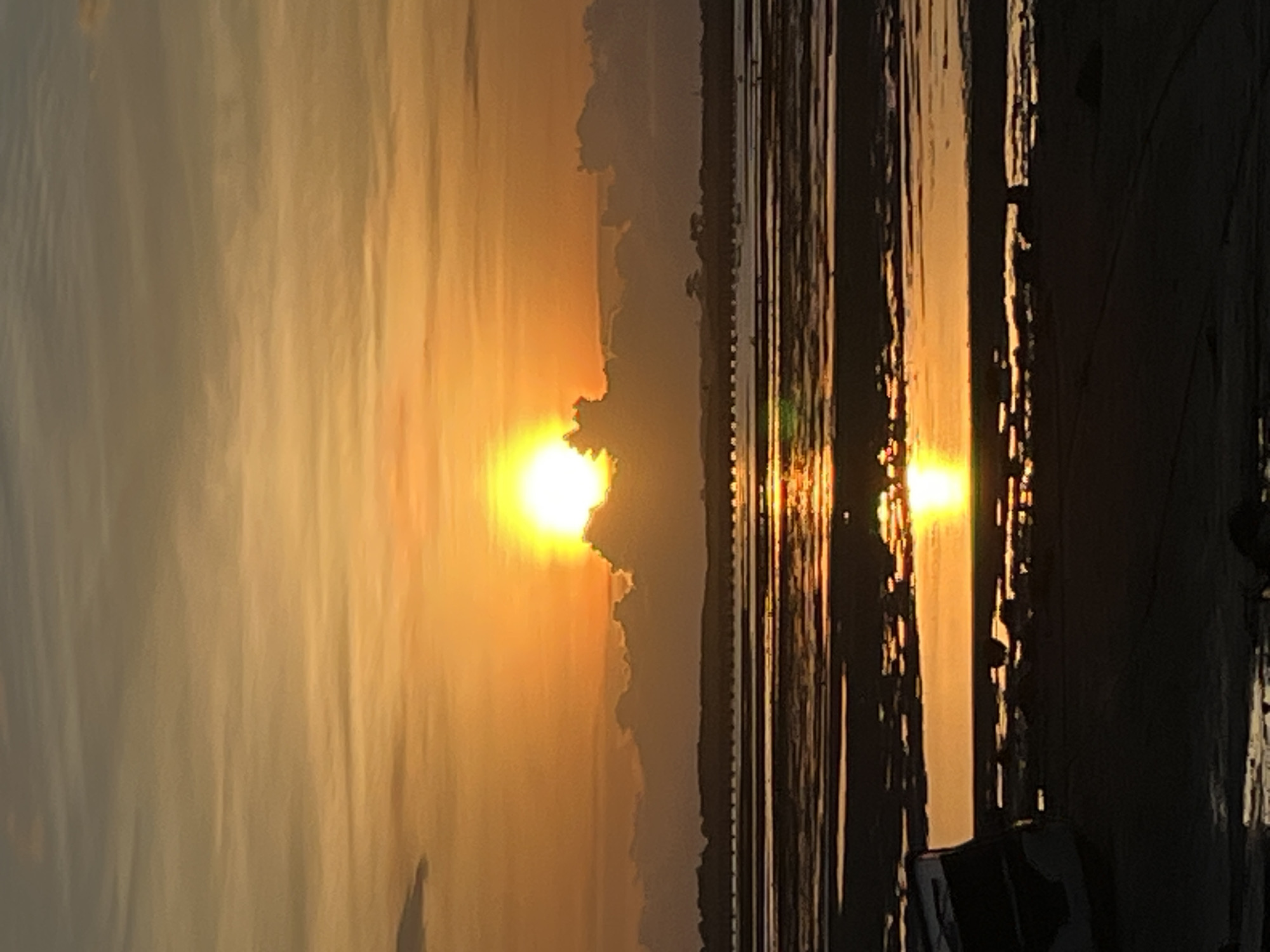 The image depicts a serene sunset over a body of water, likely a sea or large lake, in Kuta Selatan, Indonesia. The dominant colors are warm golden, orange, and yellow hues, indicating the golden hour.
The sun is a brilliant, glowing orb, partially obscured by soft, dark-edged clouds, and appears to be descending towards the horizon. Its intense light illuminates the sky with a gradient of warm colors. A distant, low-lying landmass or coastline is faintly visible along the horizon, silhouetted against the bright sky, with some atmospheric haze.
The surface of the water intensely reflects the sun's light, creating a shimmering, vertical path of bright yellow and orange that stretches from the horizon towards the foreground, suggesting ripples or gentle waves. The rest of the water surface appears darker, reflecting muted tones of the sky.
In the right foreground, several dark, indistinct vertical shapes are visible, possibly remnants of a pier, wooden poles, or other structures along the shore, casting long shadows and creating a stark contrast with the brilliant reflections. A less defined, dark object is present in the lower right corner, possibly part of a boat or a natural shoreline feature.
The overall scene conveys a sense of tranquility and natural beauty. There is no visible text in the image.