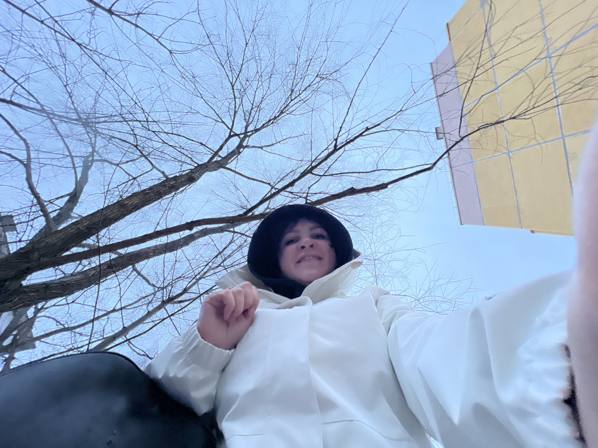 A low-angle, wide-shot photograph depicts a woman in a white jacket and dark hat, possibly a hood, against a backdrop of bare tree branches and a pale sky. A light yellow building with pink trim is visible in the upper right corner. The woman appears to be holding a camera, likely taking a selfie. Part of a dark, undefined object is in the lower left corner. The location may be in Noviy, Russia, based on prior information.