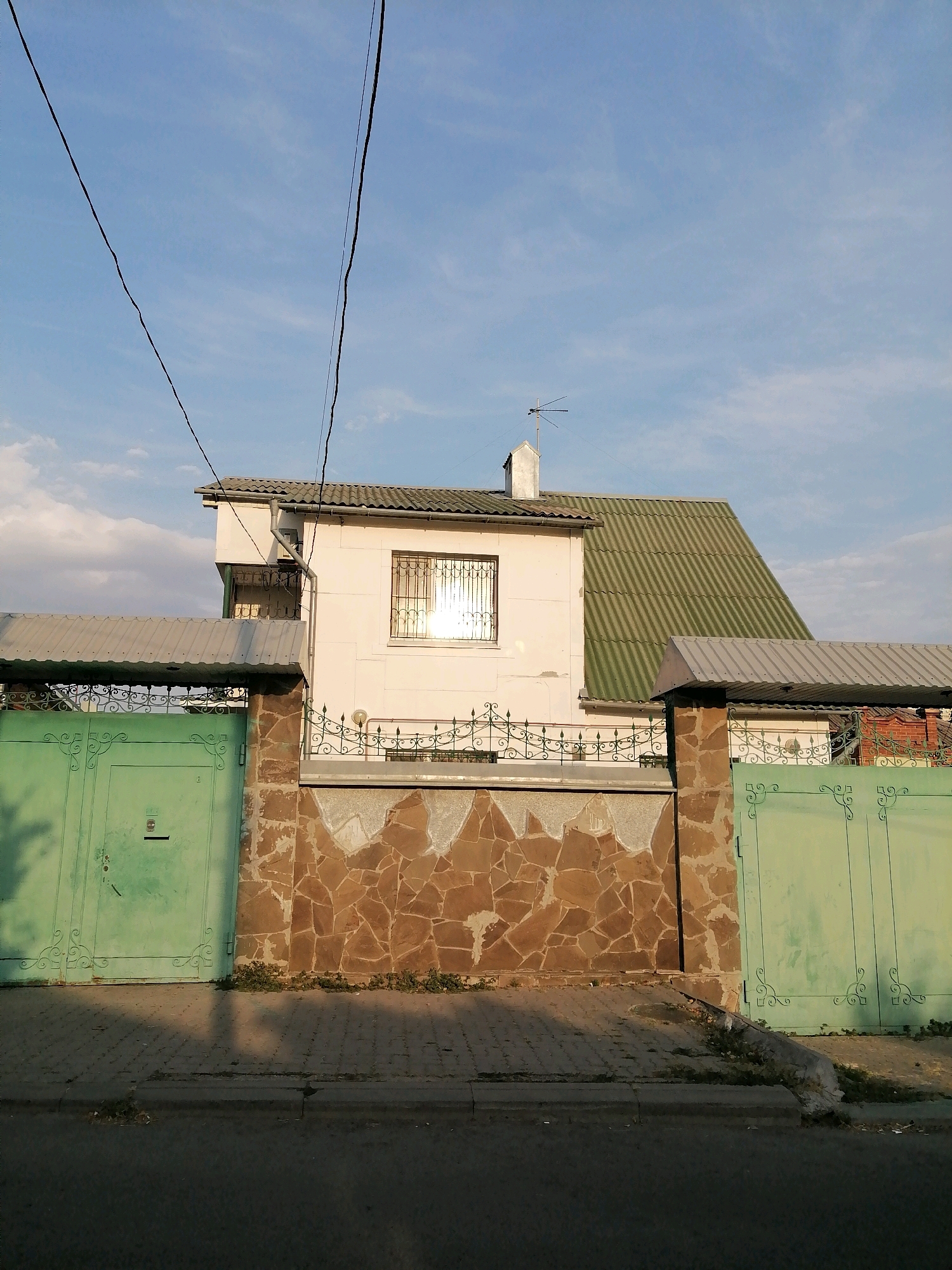 A two-story house with a green, corrugated roof is partially visible behind a stone and metal fence in Rostov-on-Don, Russia. The house has a light-colored facade, a chimney, and a window with bars. The fence features decorative green metal gates on either side of a section of stone cladding. A sidewalk runs in front of the fence, and a street is visible in the foreground. The sky is blue with scattered clouds, and power lines are visible overhead. The scene appears to be a residential street.