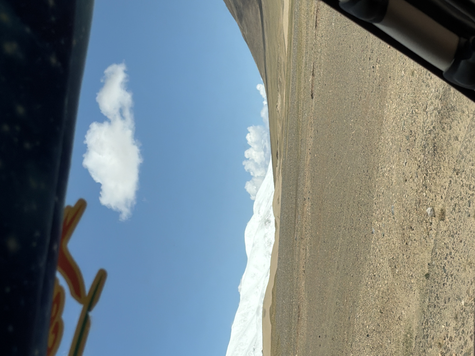 The image captures a vast, arid landscape under a clear blue sky, likely taken from inside a vehicle. In the foreground and extending to the right, there is an expanse of dry, gravelly, and rocky terrain. Dominating the mid-ground is a large, white, snow-capped or very light-colored mountain range with visible peaks and contours. The sky above is a bright blue with scattered white, puffy cumulus clouds.
On the far left side, a dark, blurred object, likely part of a vehicle's interior or structure, frames the shot. Below this object, partially visible and significantly blurred, are yellow and orange letters, possibly forming part of a sign. Another dark, blurred object, appearing to be part of the vehicle's exterior (e.g., a window frame or roof rack), is visible in the upper right corner.
The scene depicts a bright, sunny day, with strong natural light illuminating the landscape. No human or animal activity is visible. The overall setting suggests a high-altitude, mountainous, and somewhat desolate environment, consistent with areas found in regions like Labrang township, China.