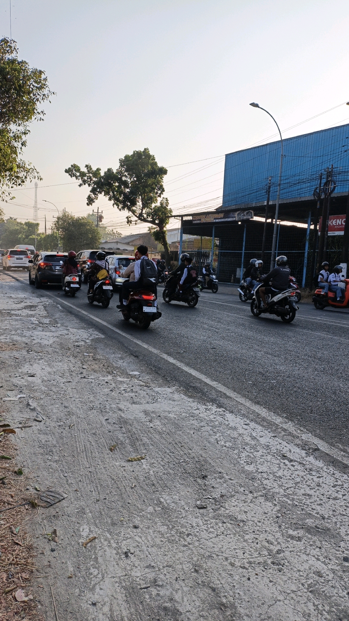 In Sumbring, Indonesia, a busy street scene unfolds under a hazy sky during daylight hours. A line of cars and motorcycles navigates the asphalt road, suggesting moderate traffic flow. The foreground of the image is dominated by a textured, greyish-brown dirt and gravel road, showing tire tracks and scattered debris, indicating a less maintained area adjacent to the main thoroughfare.
On the street, several individuals are seen riding motorcycles. One motorcycle in the center is ridden by two people, the driver wearing a backpack. To the left, a person in a red headscarf is on a scooter. Further down the road, other motorcyclists, some with helmets, are visible, indicating adherence to traffic safety. A dark-colored car and a white vehicle are also part of the procession.
The background reveals a commercial area with a large building featuring a blue, corrugated metal facade. A sign on the building reads "KENC," with some smaller text below. Utility poles and wires are present, typical of an urban environment. Trees with green foliage dot the landscape, some appearing well-established, others smaller. The overall impression is one of a typical day in a developing urban area, with transportation and commercial activity in full swing.