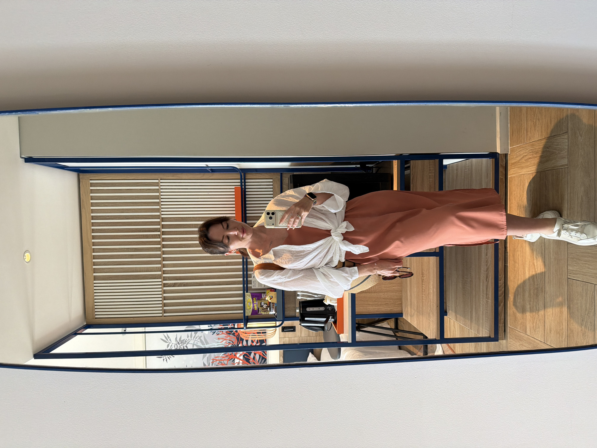 A full-length mirror reflects a woman in a hotel room. She is standing and taking a selfie with her phone. She is wearing a coral-colored midi dress, a white open button-down shirt tied at the waist, and white sneakers. She has a straw bag slung over her shoulder and is holding a pair of sunglasses in her right hand. The room has a modern design with wooden paneling on the wall and blue accents on the mirror frame and shelves. A bed is visible in the reflection behind her.