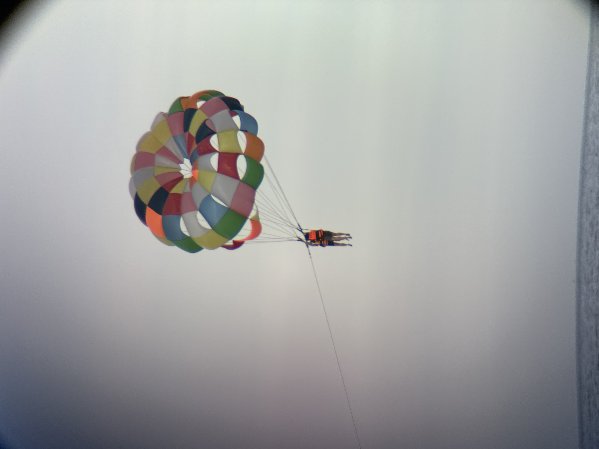 This media file captures a vibrant, multicolored parachute billowing against a hazy, overcast sky. Two individuals are visible harnessed beneath the parachute, appearing to be parasailing. Their bodies are horizontal, suggesting they are suspended in the air, experiencing the activity. The parachute itself is a prominent feature, its segments displaying a cheerful array of colors, including pink, orange, yellow, green, blue, and white, arranged in a circular pattern. The scene is dominated by the sky, which is a soft, uniform gray, indicating a diffused light, likely from the overcast conditions. A thin, dark line extends downwards from the parachute, presumably a tow rope. The right edge of the image shows a dark, textured area which may be a distant landmass or structure, and a faint line of water or the sea. The overall impression is one of leisure and adventure, with the bright parachute offering a pop of color against the muted backdrop. The time of day is indeterminate due to the overcast sky, but the soft light suggests it is not the harsh glare of midday. The setting appears to be a coastal area, given the presence of what might be the sea. There is no visible text in the image.