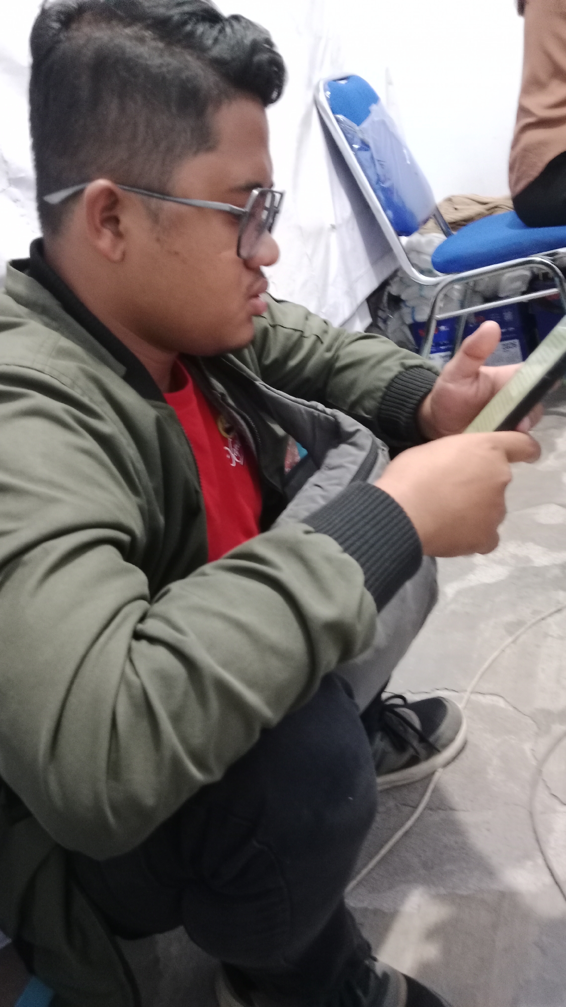 A young man with glasses and short, styled dark hair is seated, looking at his phone, which he holds in his right hand. He is wearing a green bomber jacket over a red t-shirt and dark pants. The background shows a white wall and blue chairs, suggesting an indoor setting. The floor is a light-colored, textured surface. There are no immediate indications of this scene being in Blitar, Indonesia.