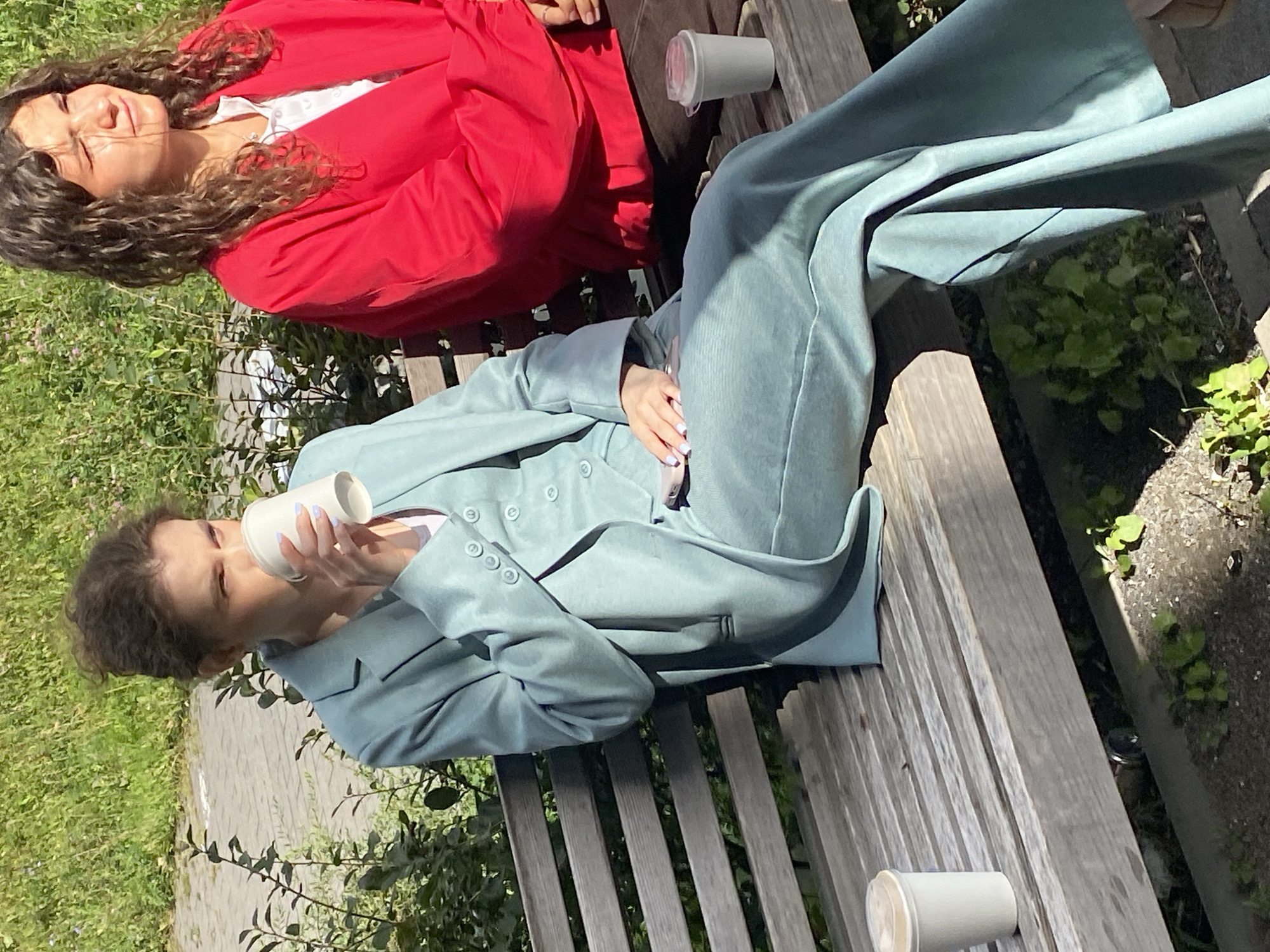 Two individuals are seated on a wooden park bench outdoors. The person on the left is wearing a teal suit and drinking from a white paper cup. The person on the right is wearing a bright red jacket and a white shirt. The background consists of green foliage and a paved area. The scene appears to be in a park or public outdoor space.