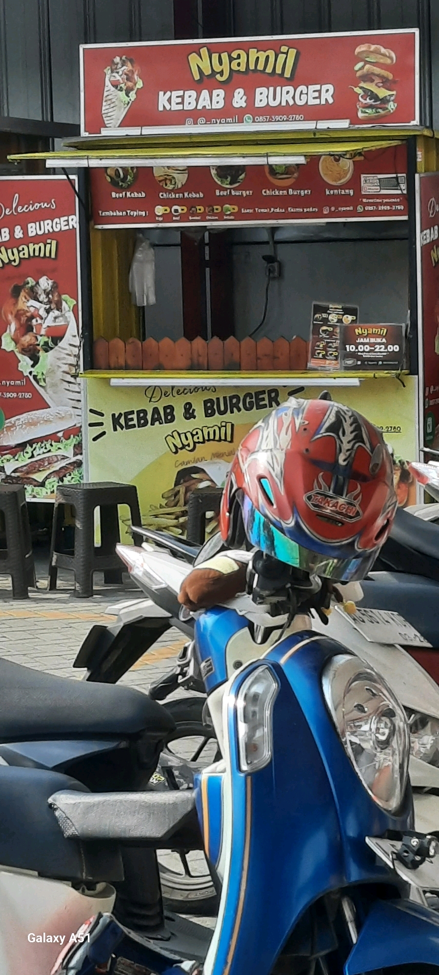 This is a medium shot, eye-level, outdoor photograph of a food stall in Wlingi, Indonesia. The stall, named "Nyamil Kebab & Burger," is adorned with vibrant red and yellow signage showcasing images of kebabs and burgers. The stall is set up on a street with a cobblestone pavement visible in the foreground. A motorcycle with a blue and white body is parked in front of the stall, with a red helmet with a reflective visor resting on its handlebars. The helmet has a Japanese design with flames and the brand "TAKAGHI" visible. Behind the stall, a glimpse of a dark-colored wall and an electrical outlet can be seen. The overall scene suggests a bustling street food vendor ready to serve customers.