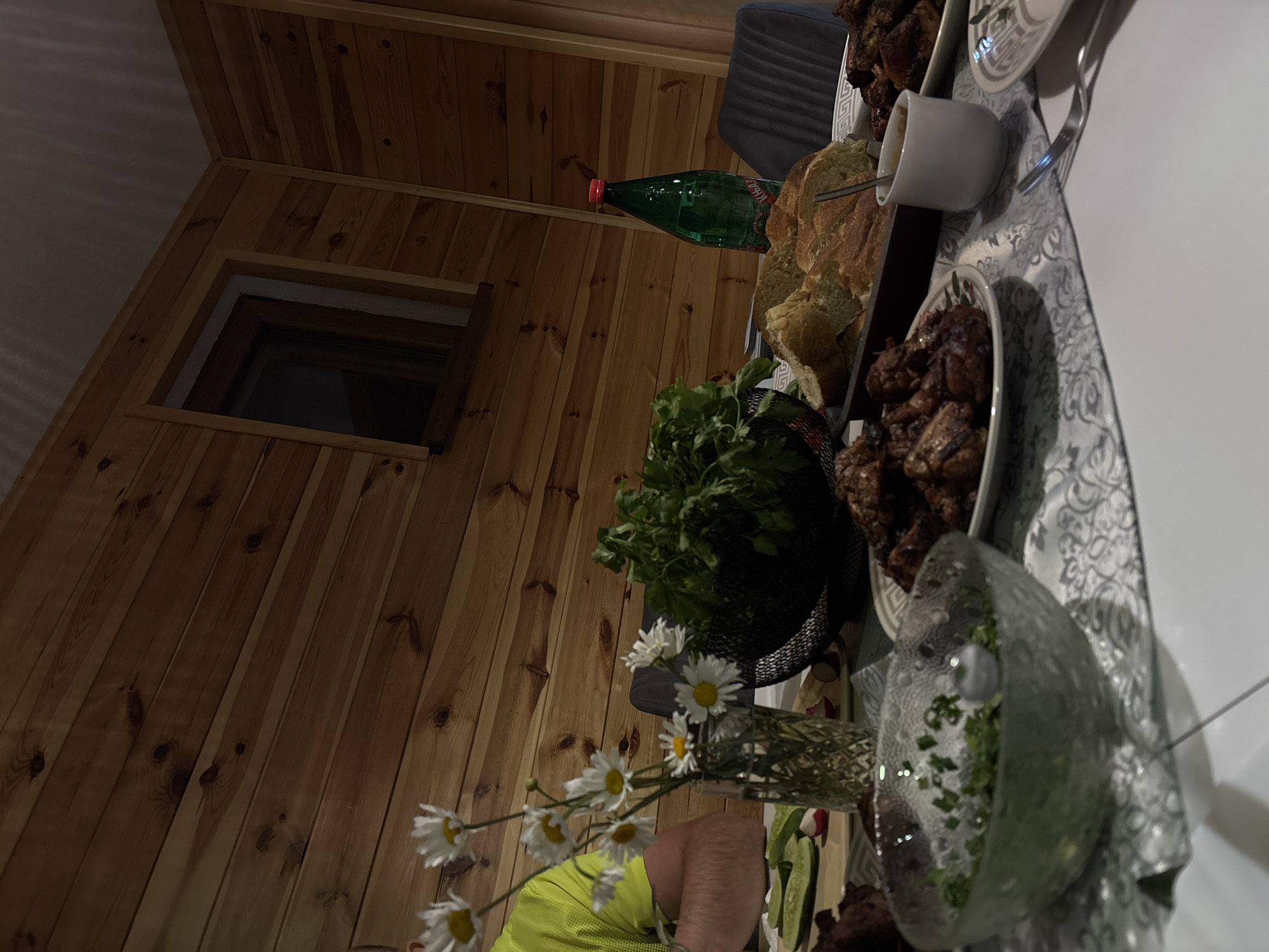 The image depicts an outdoor dining scene, likely a meal enjoyed on a patio or balcony, as indicated by the wooden paneled walls and natural lighting. The setting appears to be in Virak, Montenegro, given the context. On a table, laid with a patterned tablecloth, are several dishes of food. In the foreground, a glass bowl contains a green salad with chopped herbs. To the right, a white ceramic cup holds a sauce or dip. Further to the right, plates with cooked meat, possibly chicken or pork, are visible. In the center of the table, a dark rectangular tray holds slices of bread. Behind the food, a green bottle of water with a red cap stands upright. A bunch of fresh green herbs is placed in a dark basket. To the left, a vase holds a small bouquet of daisies. A person wearing a bright yellow t-shirt is partially visible on the left side of the frame, with their arm resting on the table. The overall atmosphere suggests a casual meal shared with company.