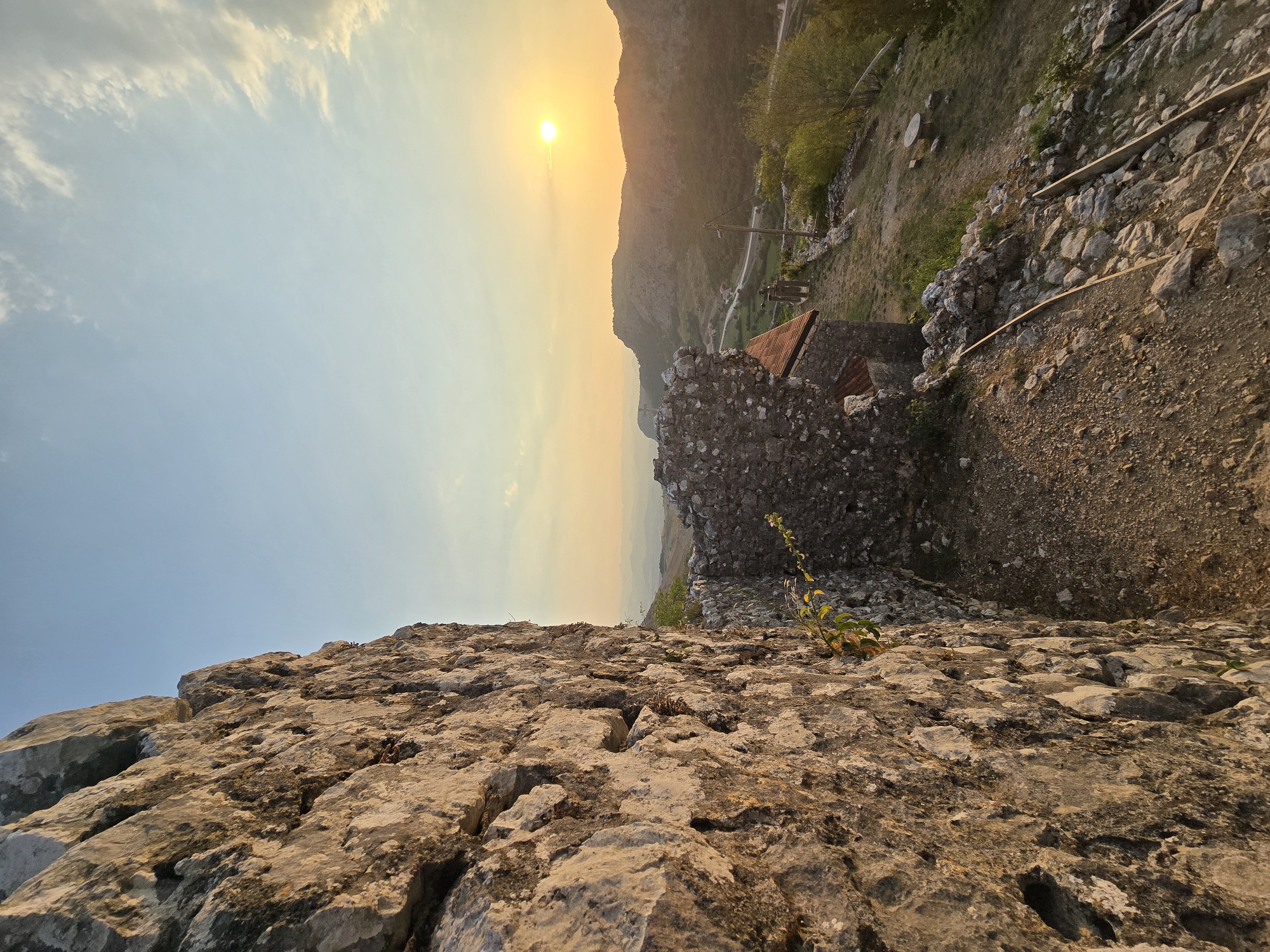 The image depicts a sunset view from the walls of Medun Fortress in Medun, Montenegro. The foreground is dominated by the rough, textured stone of the fortress wall, which occupies the left side of the frame. Beyond the wall, a partially ruined stone structure with a red-tiled roof is visible. The middle ground shows a sloping, overgrown terrain with scattered rocks and some wooden planks. In the background, a valley unfolds with a road winding through it, bordered by hills and mountains. The sun, a bright orb, is low on the horizon, casting a warm, golden light over the landscape and creating a soft glow in the sky, which transitions from a clear blue to hues of orange and yellow near the sun. There are no people or animals present in the image. The scene conveys a sense of historical ruin and natural beauty.