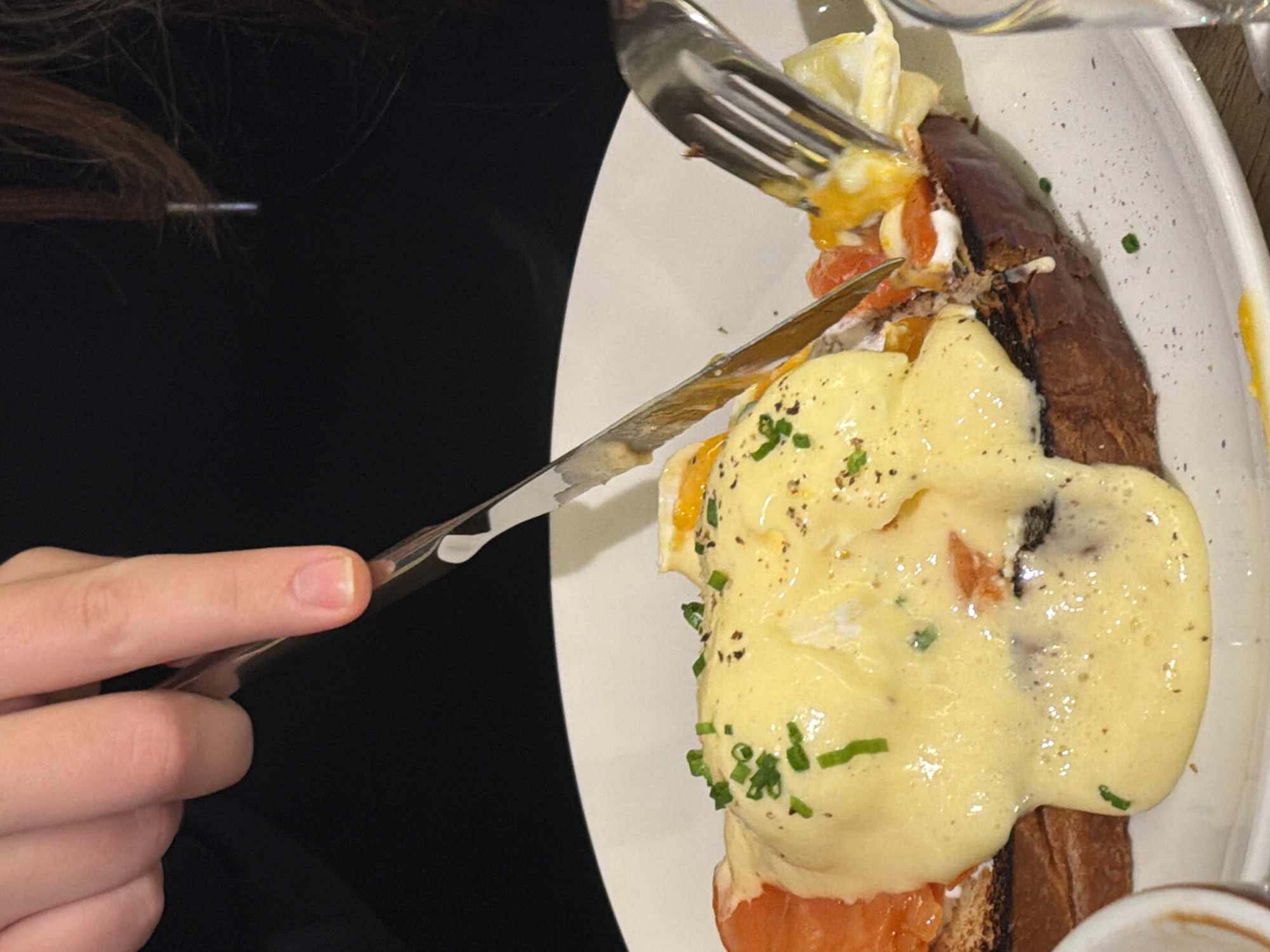 A person's hand holds a knife, cutting into a dish of Eggs Benedict with salmon on a white plate. A fork is also in use. The location is unknown; Belgrade, Serbia is not identifiable.