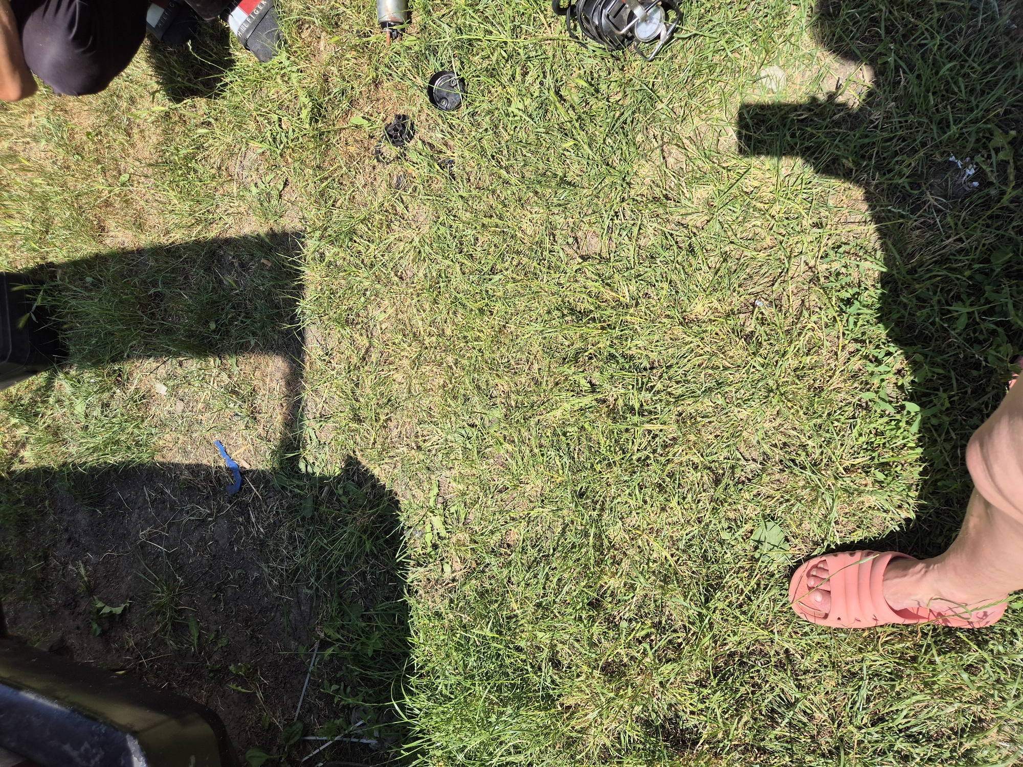 The image shows a ground-level view of a grassy area with patches of dirt, suggesting an outdoor setting. The sunlight indicates it is daytime, likely a sunny day given the clear shadows.
Several objects are scattered on the ground. In the top left, part of a person's leg wearing black pants and black shoes with white and red stripes is visible. To the right of this, there are fishing equipment items, including a fishing reel and what appears to be a fishing rod holder or part of a reel's base. There is also a dark circular object, possibly a cap or a small container, and a few other unidentifiable dark items. A blue, curved object, possibly a piece of plastic or tubing, lies on the dirt in the lower left quadrant.
In the bottom right corner, a person's bare foot wearing a pink slide sandal is visible, resting on the grass. The presence of people and fishing gear suggests an outdoor recreational activity. The ground is a mix of green grass and dry, bare soil. The shadows cast by an unseen person or object are prominent on the grass.
There is no visible text in the image. The overall scene evokes a casual, outdoor setting in what appears to be Ukraine, possibly during a fishing trip or a break in outdoor activities.