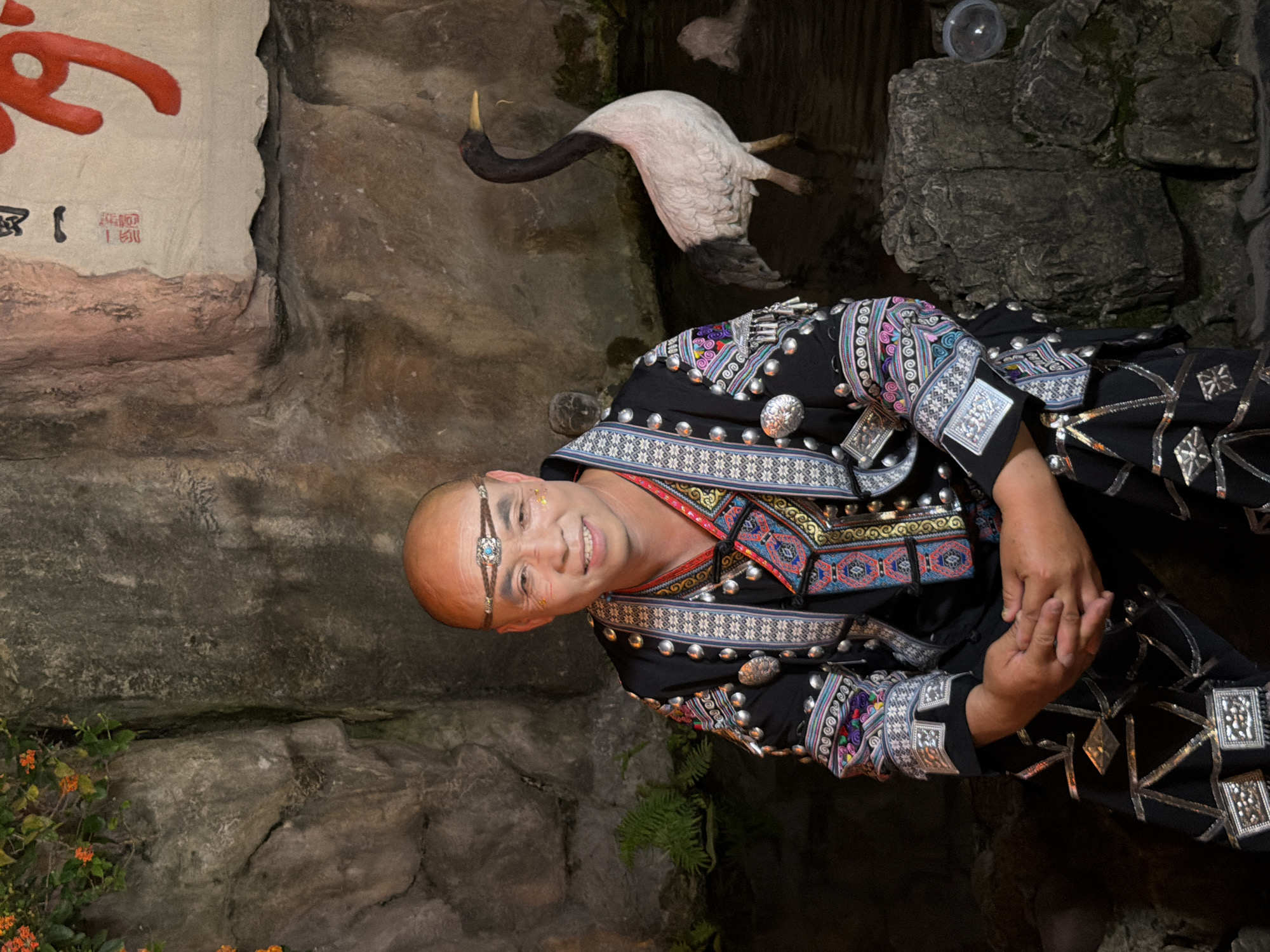 In a dimly lit, rocky cavern, a man adorned in a black, intricately embroidered traditional costume with silver embellishments smiles at the camera. The costume features colorful geometric patterns along the neckline and cuffs. He wears a delicate silver headband with a blue gemstone over his forehead and has subtle glitter applied to his cheeks. His hands are clasped in front of him. To his right, a taxidermied crane with black wings and a white body is poised in mid-flight, its neck extended. The background consists of rough, textured rock formations, with some greenery and small orange flowers visible in the lower left corner. A red Chinese character is painted on a white surface on the upper left side of the image, partially obscured by the rock. The scene appears to be an indoor theatrical or cultural performance, given the costuming and staged elements.