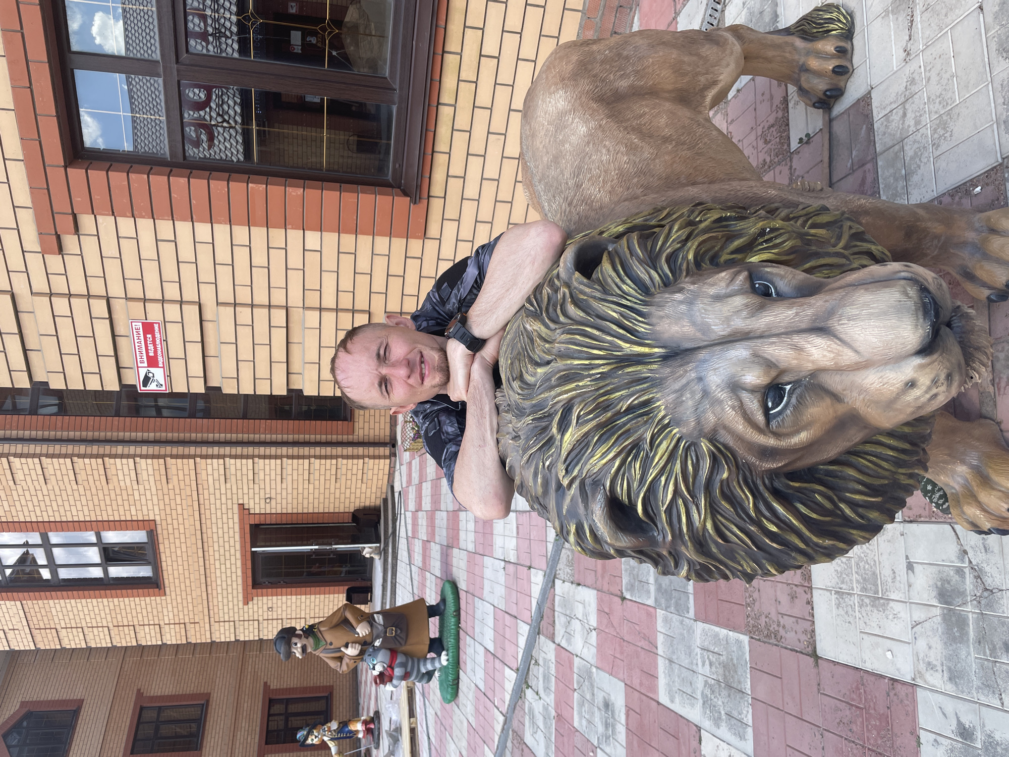 A man straddles a lion statue in Sergeevka, Russia. The man, fair-skinned with short hair and wearing a dark patterned shirt, is positioned with his arms around the lion's head. The lion is a large, realistic-looking statue with a golden mane. The background shows a brick building with windows, and other statues are visible along the patterned tile ground. A warning sign is affixed to the brick building.