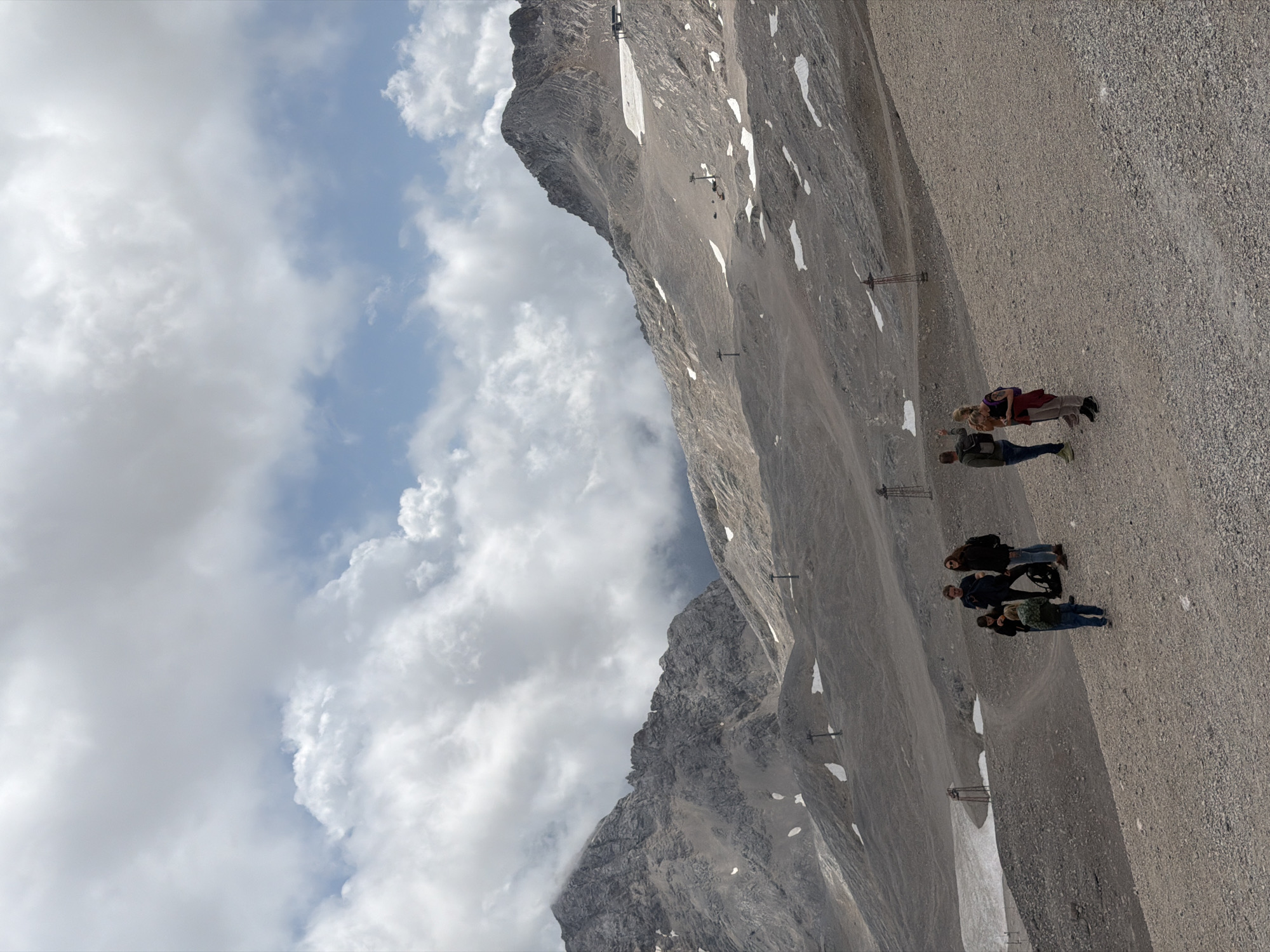 The image captures a group of five people standing on a rocky, gravelly mountain slope with scattered patches of snow. The group consists of three adults and two children. One adult, a male, is wearing a dark jacket and jeans and has a backpack. Another adult, a female, is wearing a dark jacket and jeans, and has long brown hair and sunglasses. A third adult, a female, has dark curly hair and is wearing a backpack. One child, a boy, is wearing a green jacket and blue jeans and is facing away from the camera. Another child, a girl, is wearing a pink jacket and dark pants and is holding a cell phone. In the background, there are rugged, rocky mountains with some snow-covered areas. Several ski lift poles are visible on the slopes, suggesting this is a ski resort or a mountain recreation area. The sky is partly cloudy with a mix of white and gray clouds and patches of blue sky. This scene is likely at the Zugspitze, Germany's highest peak, located near Garmisch-Partenkirchen.